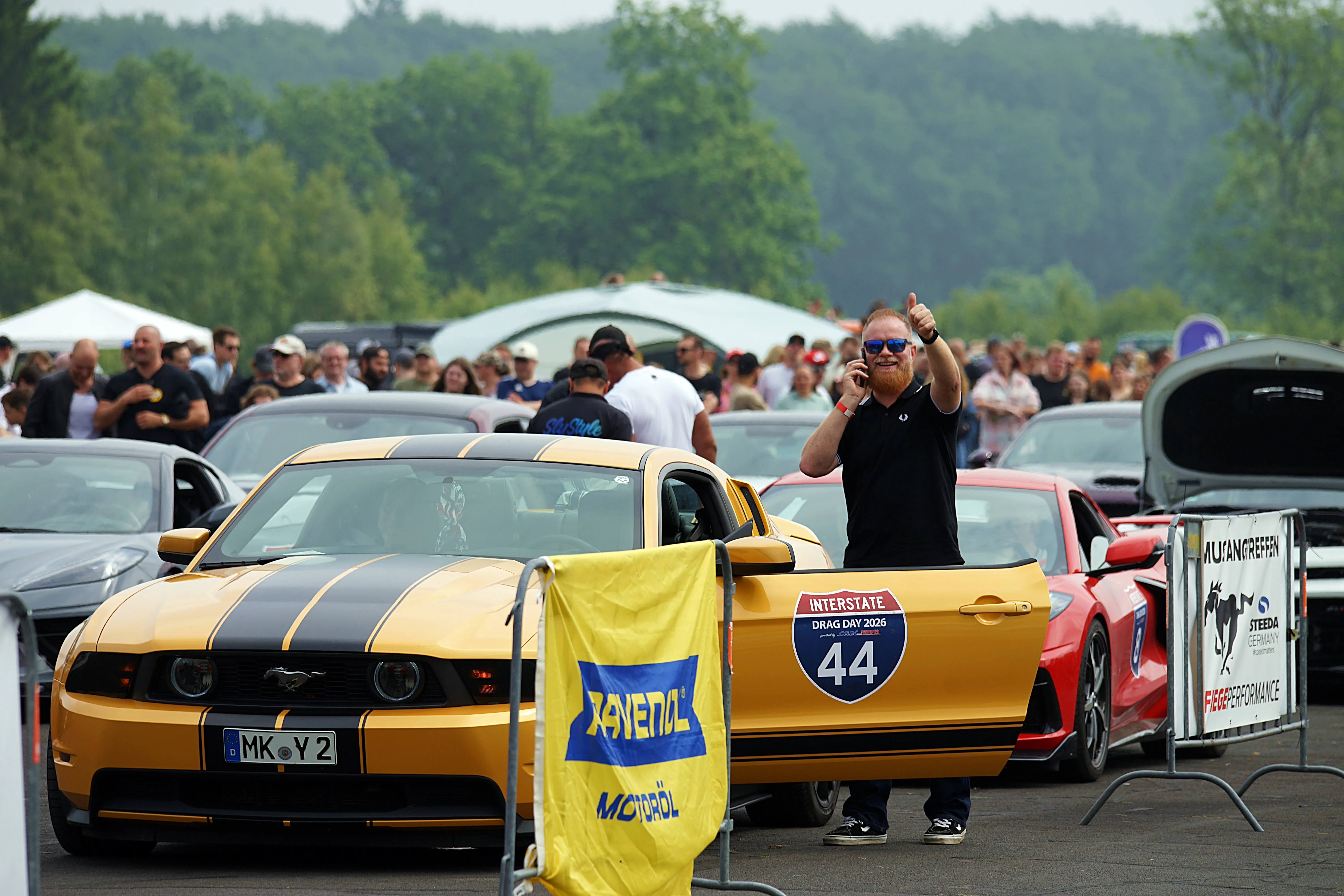 Ein Mann steht auf dem Dach eines gelb-schwarzen Ford Mustang und zeigt den Daumen nach oben. Im Hintergrund sind weitere Autos und eine Menschenmenge zu sehen.