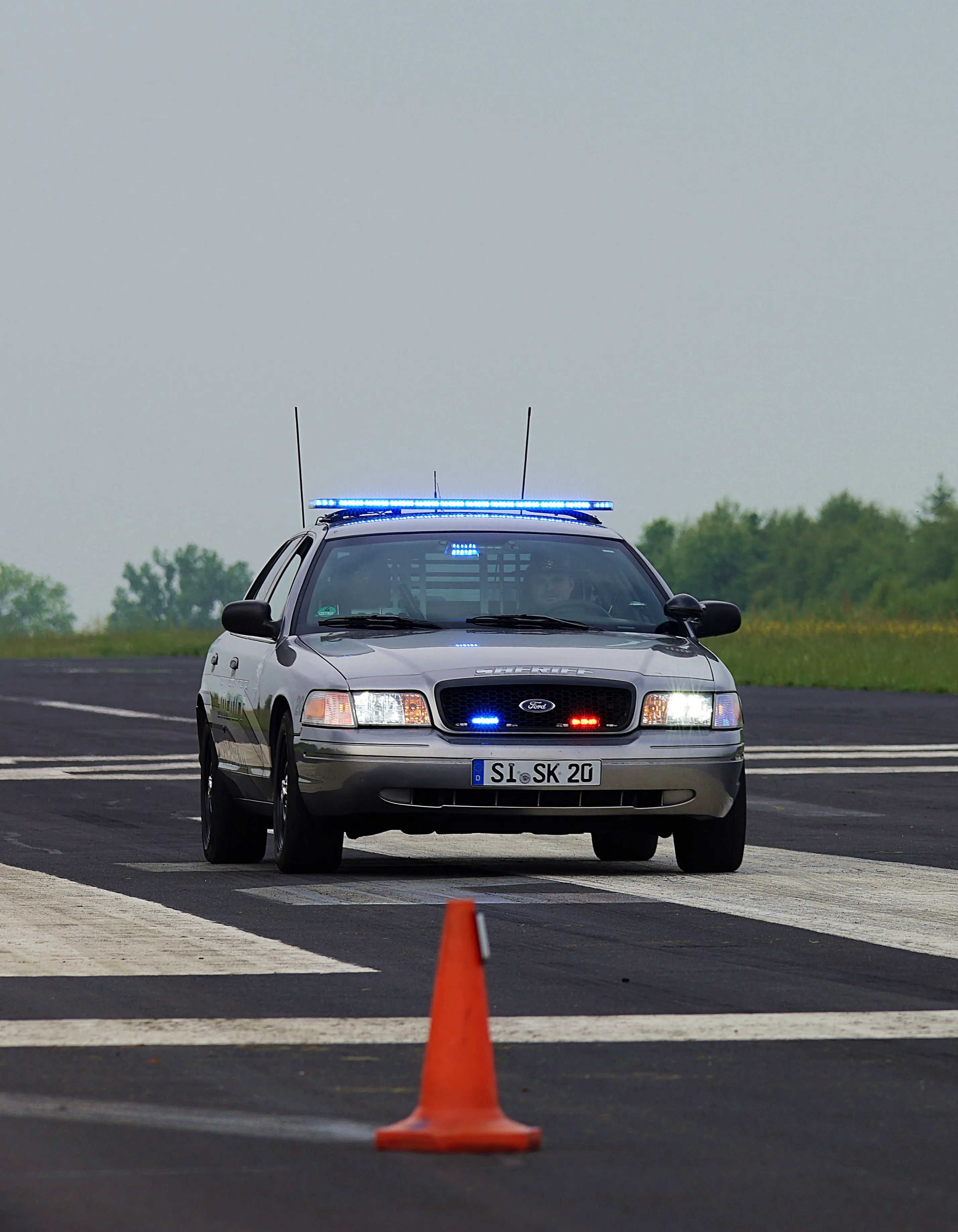 Silberner Ford Police Interceptor Cruiser mit eingeschalteten Blaulichtern auf einer asphaltierten Straße, vor dem Fahrzeug steht eine orangefarbene Verkehrskegel.