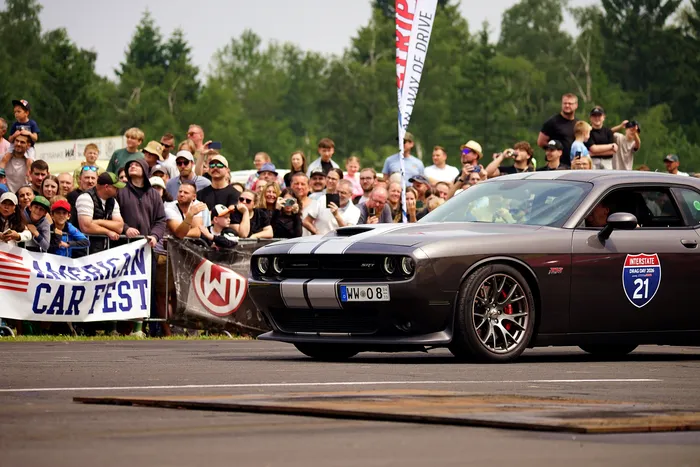 Ein dunkelgrauer Dodge Challenger fährt auf einer Straße vor einer jubelnden Menschenmenge. Im Hintergrund Banner mit der Aufschrift „American Car Fest“ und ein Schild mit der Aufschrift „Interstate Drive 21“.