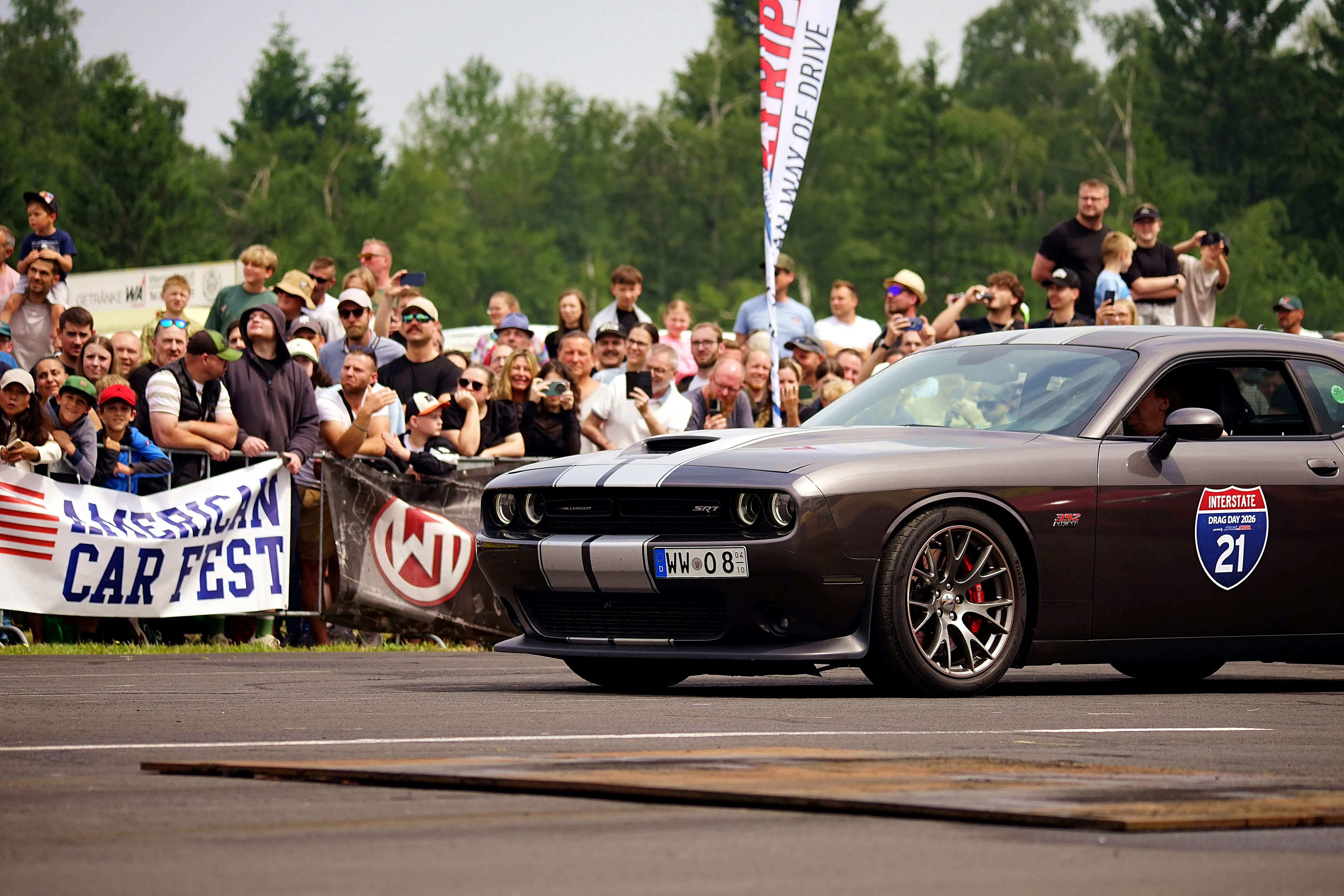 Ein dunkelgrauer Dodge Challenger fährt auf einer Straße vor einer jubelnden Menschenmenge. Im Hintergrund Banner mit der Aufschrift „American Car Fest“ und ein Schild mit der Aufschrift „Interstate Drive 21“.