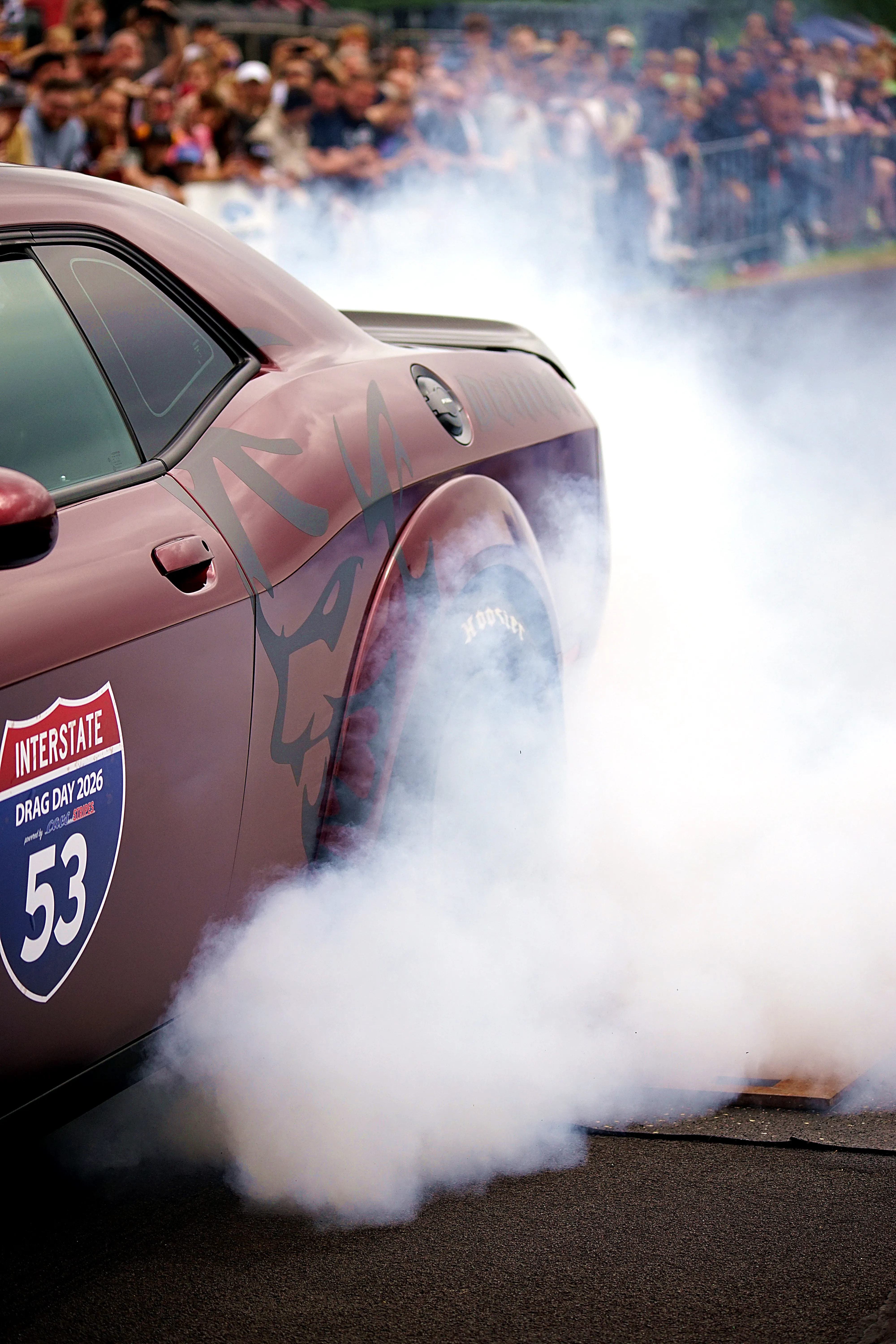 Ein rotes Muscle-Car mit der Startnummer 53 auf der Tür erzeugt dichten Rauch beim Beschleunigen, im Hintergrund eine Zuschauermenge.