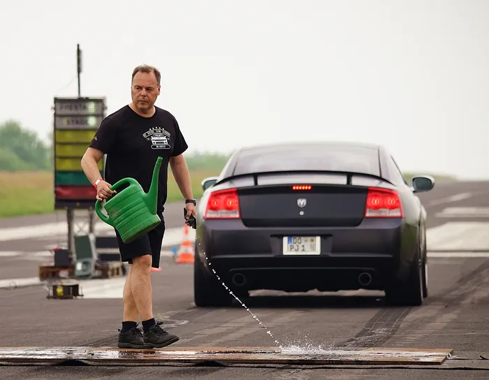 Ein Mann gießt Wasser aus einer grünen Gießkanne auf den Asphalt vor einem grauen Dodge Charger auf einer Rennstrecke.