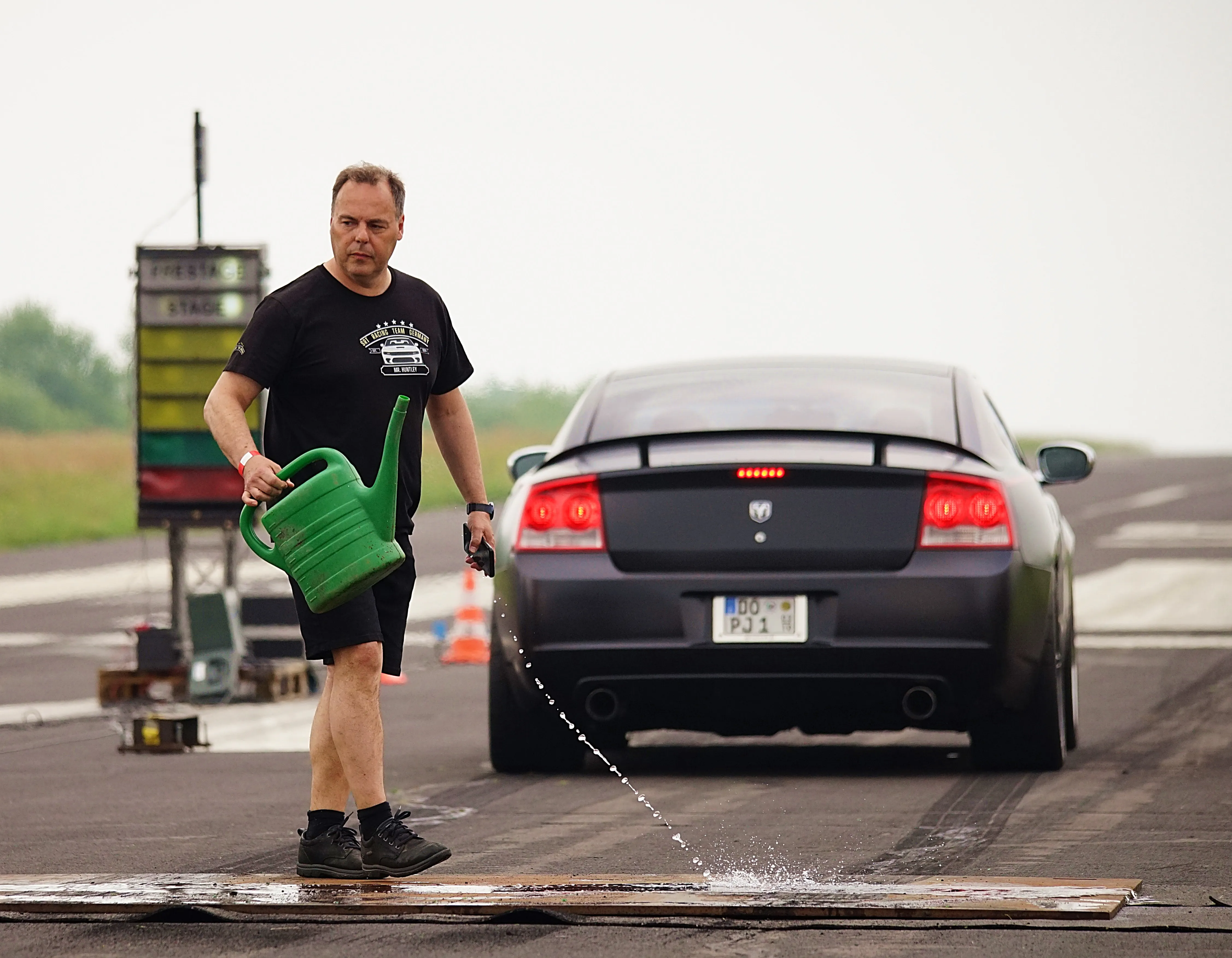 Ein Mann gießt Wasser aus einer grünen Gießkanne auf den Asphalt vor einem grauen Dodge Charger auf einer Rennstrecke.