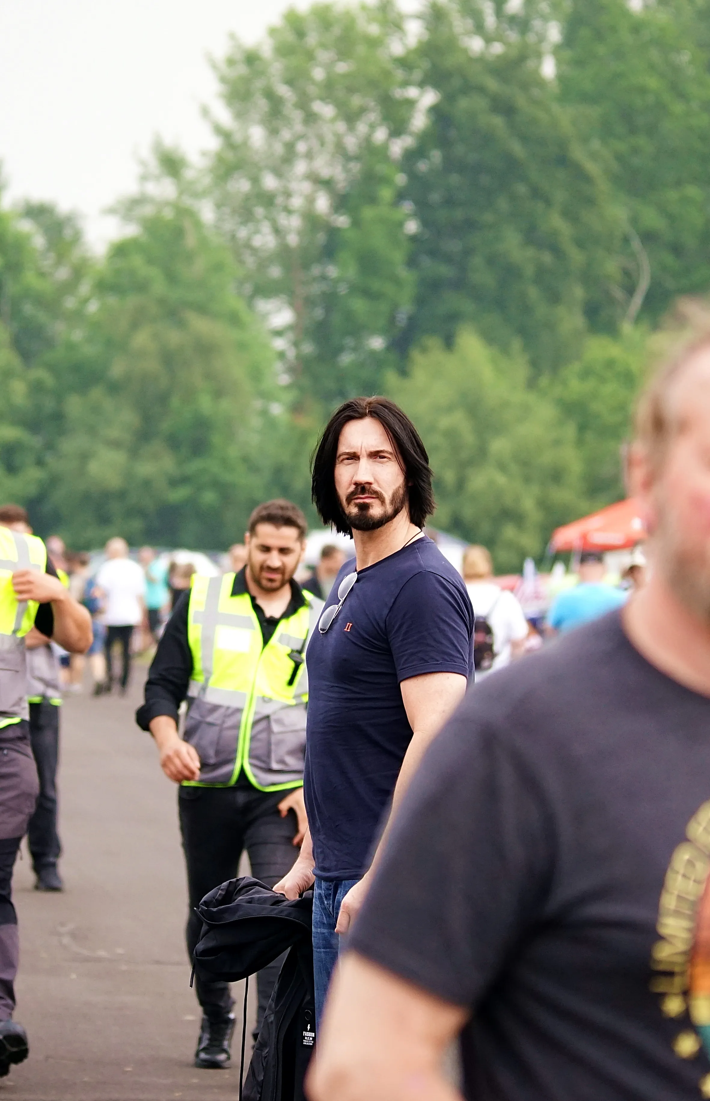 Keanu Reeves mit Bart und dunklem T-Shirt steht vor einer Menschenmenge, im Hintergrund Sicherheitskräfte mit Warnwesten.