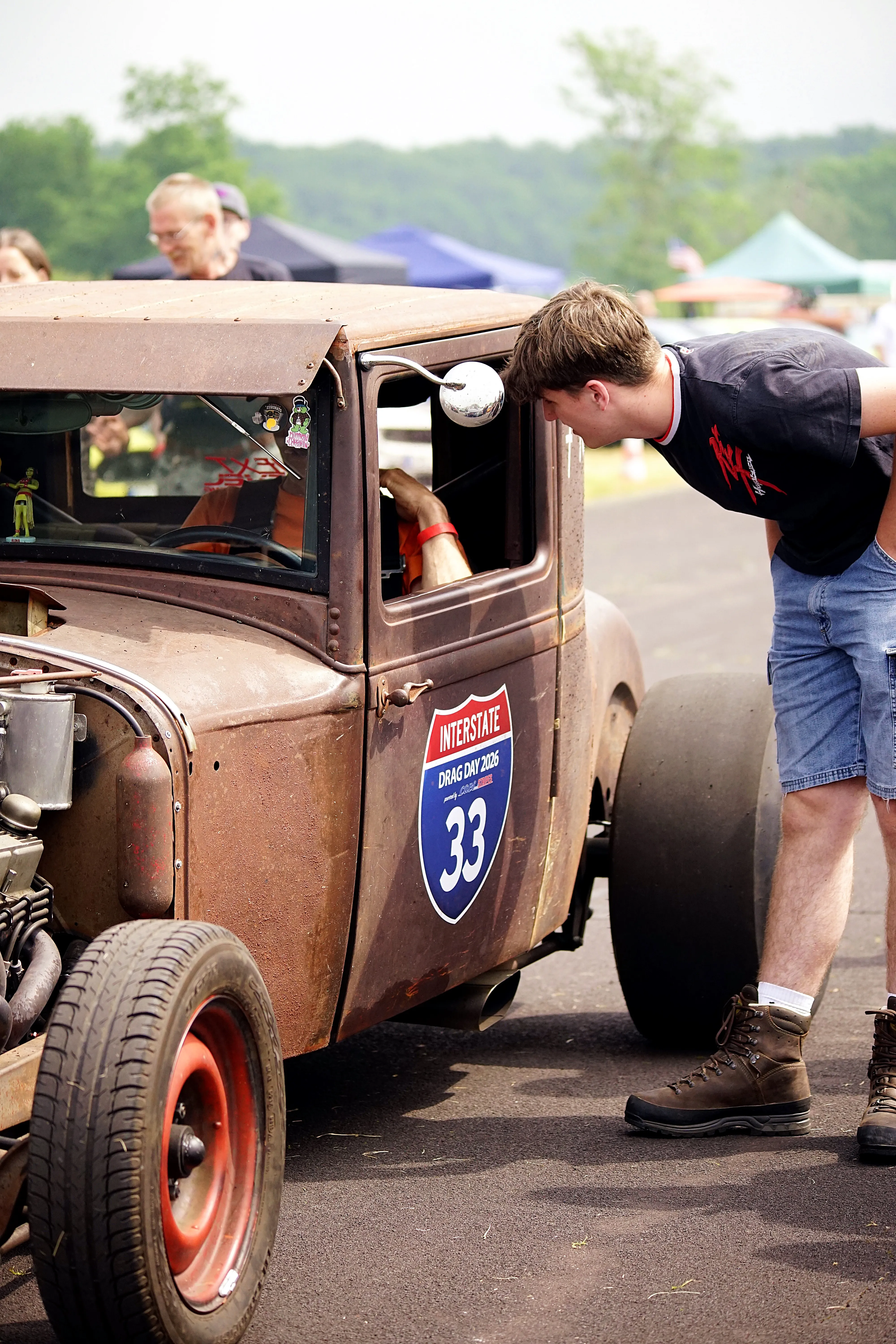 Ein Mann beugt sich über die offene Fahrertür eines rostigen, alten Hot Rods mit der Startnummer 33. Im Hintergrund ist ein weiterer Mann und ein Festzelt zu sehen.