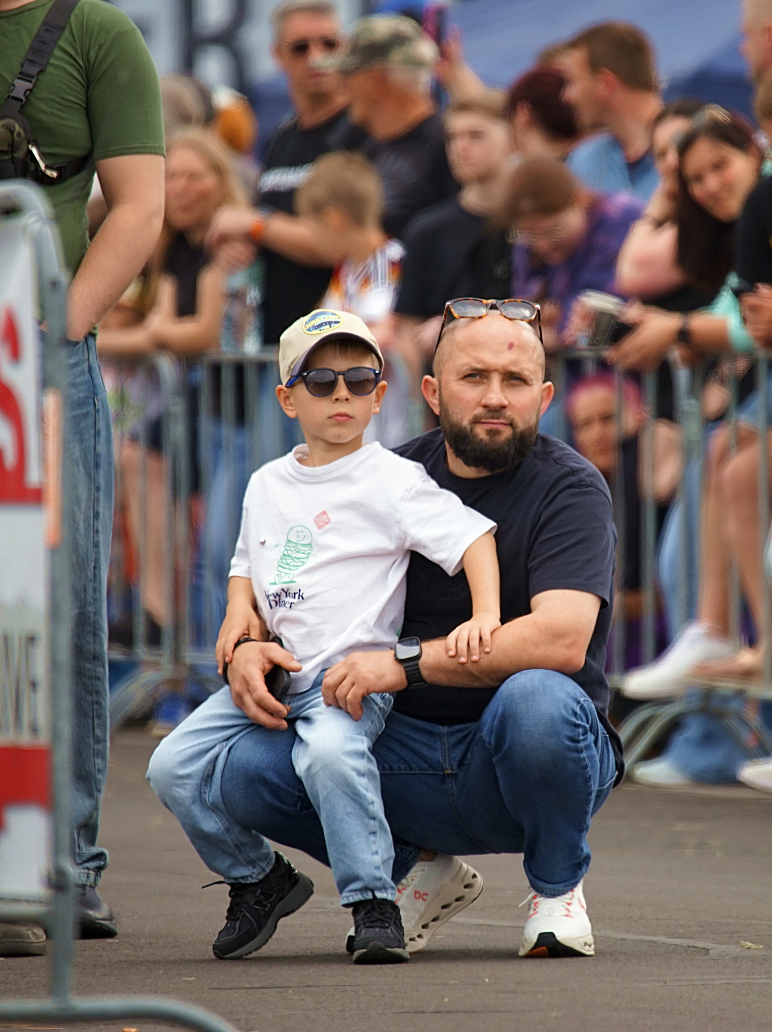 Ein Mann hockt vor seinem Sohn und trägt ihn auf seinen Schultern, während sie eine Veranstaltung mit vielen Menschen im Hintergrund besuchen.