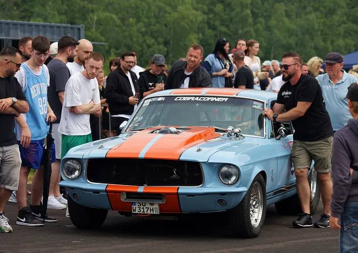 Ein hellblauer Ford Mustang mit orangefarbenen Streifen steht im Mittelpunkt einer Menschenmenge auf einer Veranstaltung. Ein Mann in Schwarz steht neben dem Fahrzeug und betrachtet es.