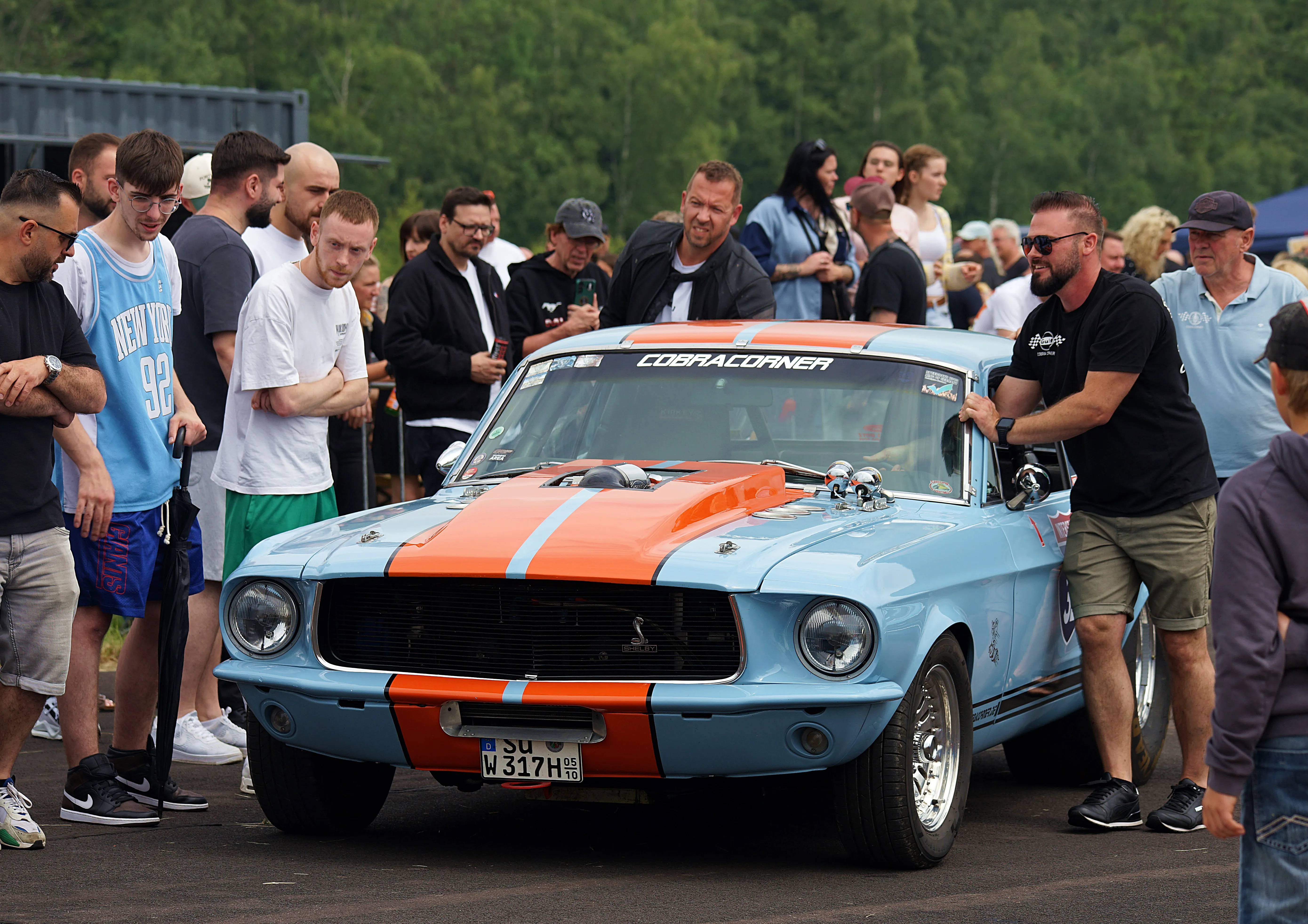 Ein hellblauer Ford Mustang mit orangefarbenen Streifen steht im Mittelpunkt einer Menschenmenge auf einer Veranstaltung. Ein Mann in Schwarz steht neben dem Fahrzeug und betrachtet es.