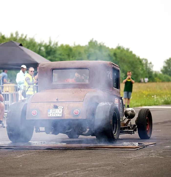 Rostiger, dunkler Hot Rod der 1930er Jahre mit rauchenden Reifen auf einer Rennstrecke, umgeben von Zuschauern.
