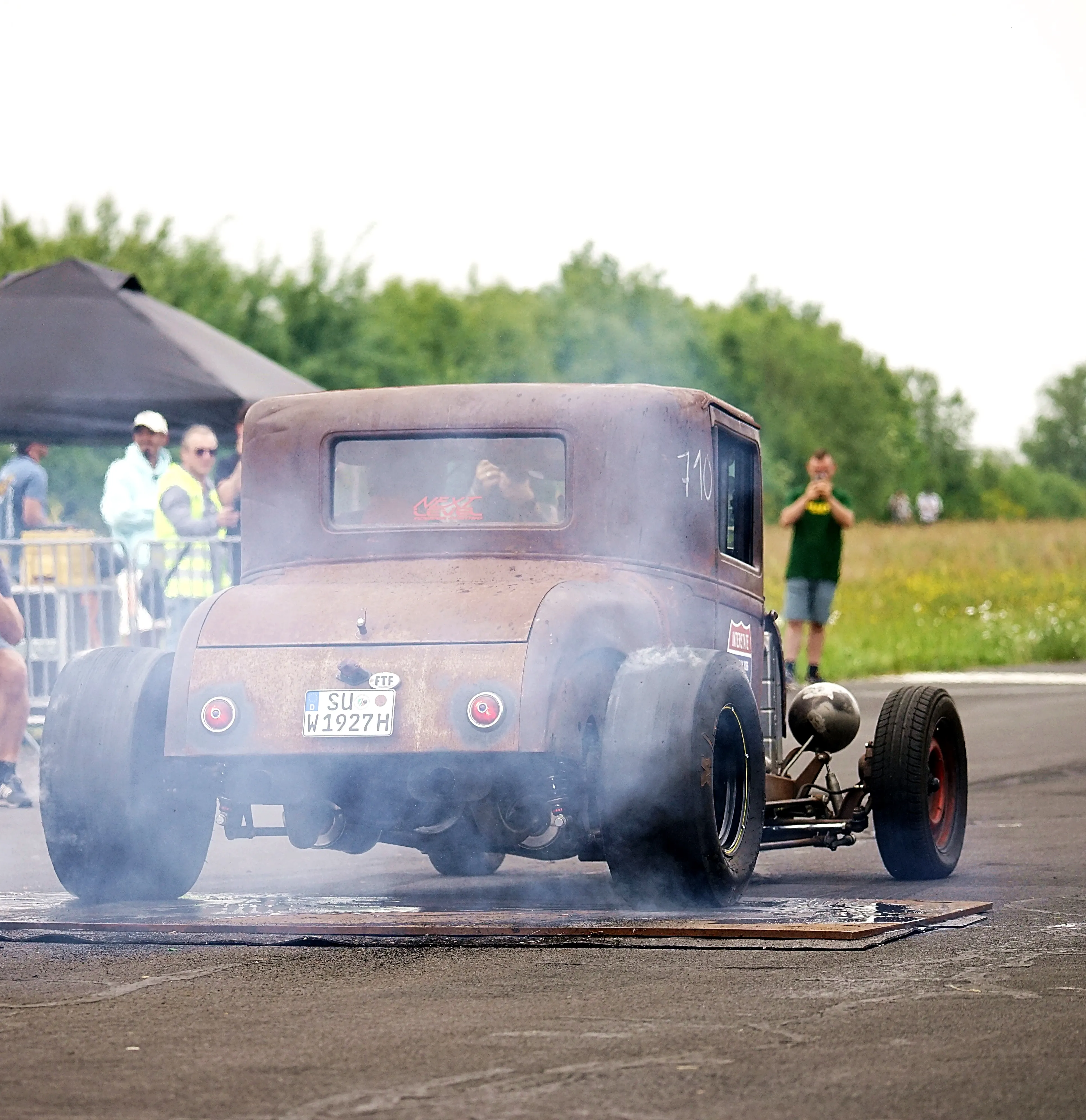 Rostiger, dunkler Hot Rod der 1930er Jahre mit rauchenden Reifen auf einer Rennstrecke, umgeben von Zuschauern.