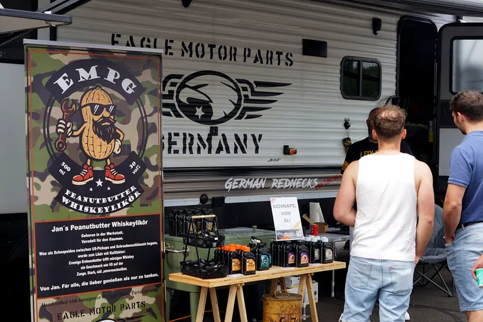 Ein Wohnwagen mit der Aufschrift „Eagle Motor Parts Germany“ und ein Stand mit Flaschen Peanutbutter Whiskeylikör und Ölprodukten.
