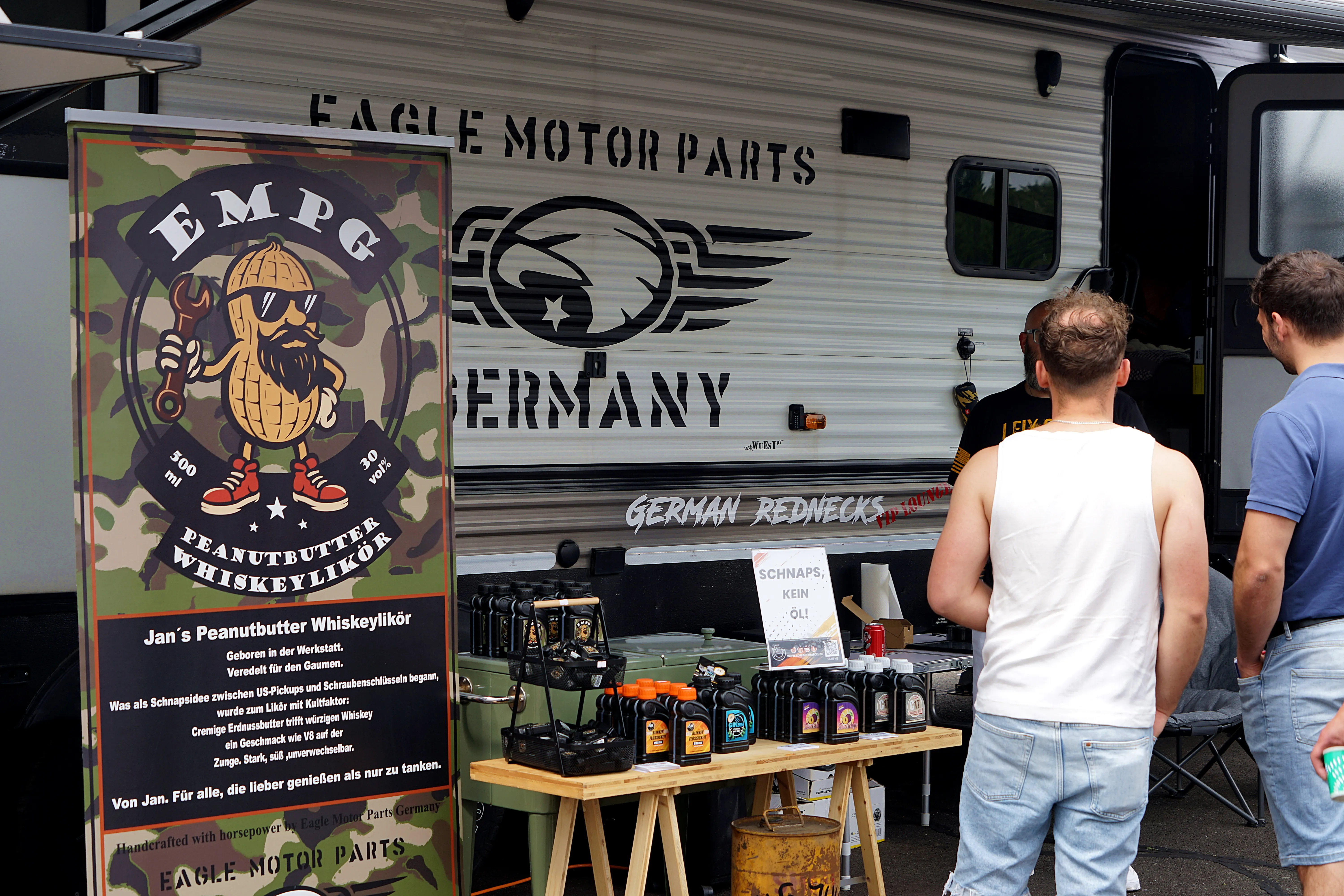 Ein Wohnwagen mit der Aufschrift „Eagle Motor Parts Germany“ und ein Stand mit Flaschen Peanutbutter Whiskeylikör und Ölprodukten.