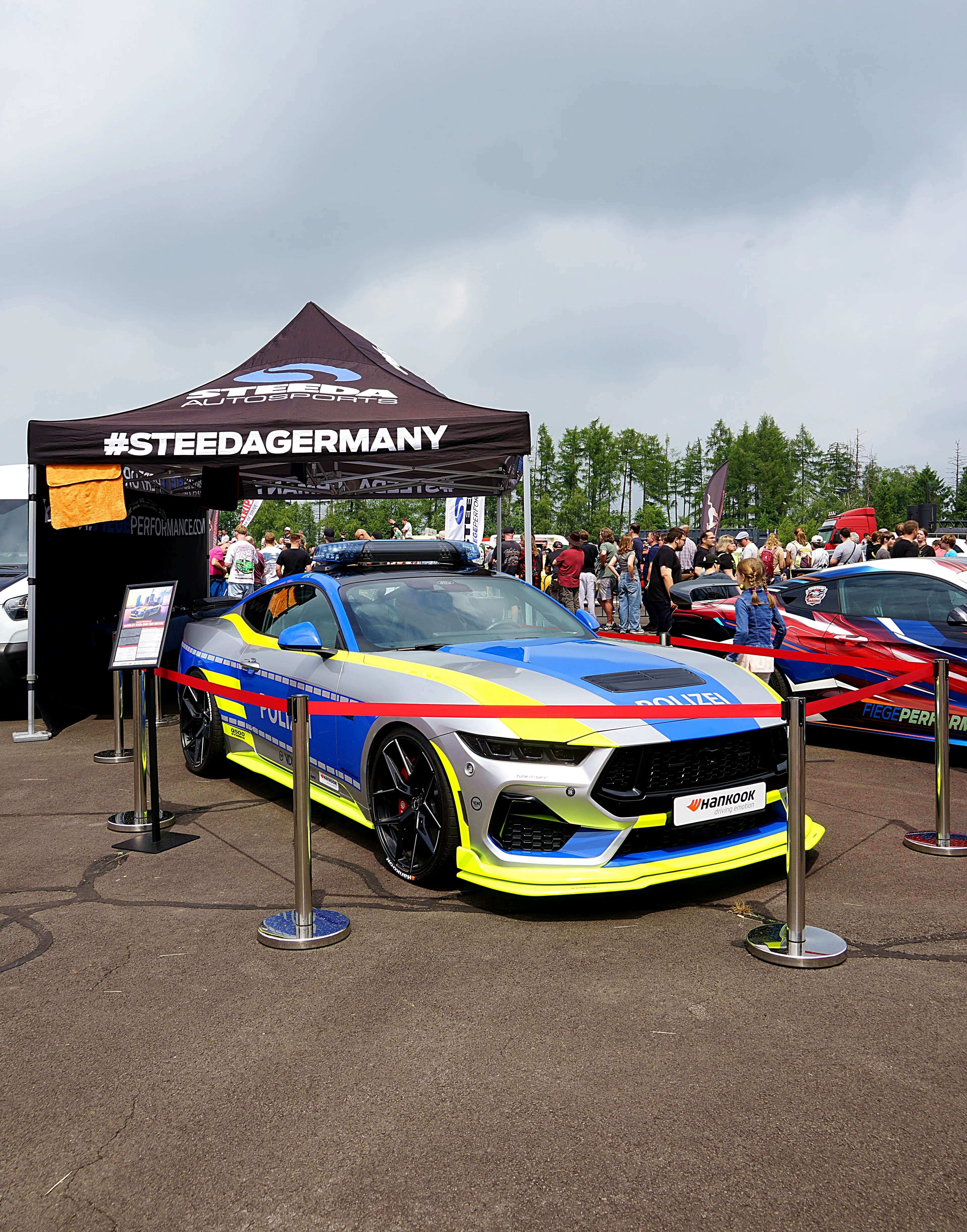 Ein Polizei-Sportwagen im auffälligen Farbdesign steht vor einem Stand mit dem Schriftzug #STEEDGERMANY. Im Hintergrund sind weitere Fahrzeuge und Besucher zu sehen.