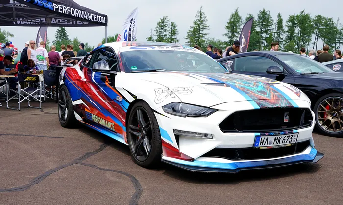 Weißer Ford Mustang mit farbenfroher Folierung und Rennstrecken-Optik steht auf einem Parkplatz, Kennzeichen WA-MK 673.