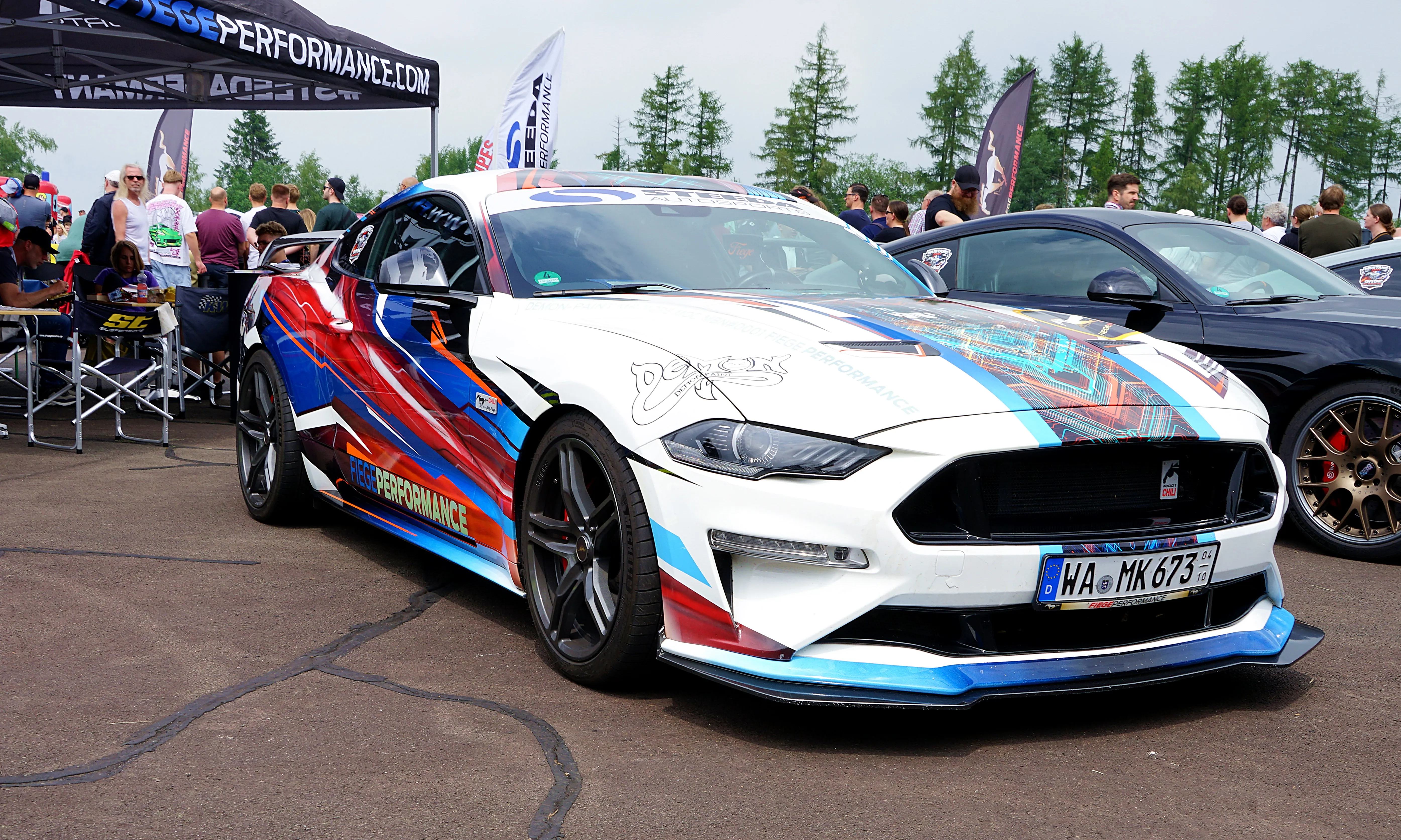 Weißer Ford Mustang mit farbenfroher Folierung und Rennstrecken-Optik steht auf einem Parkplatz, Kennzeichen WA-MK 673.