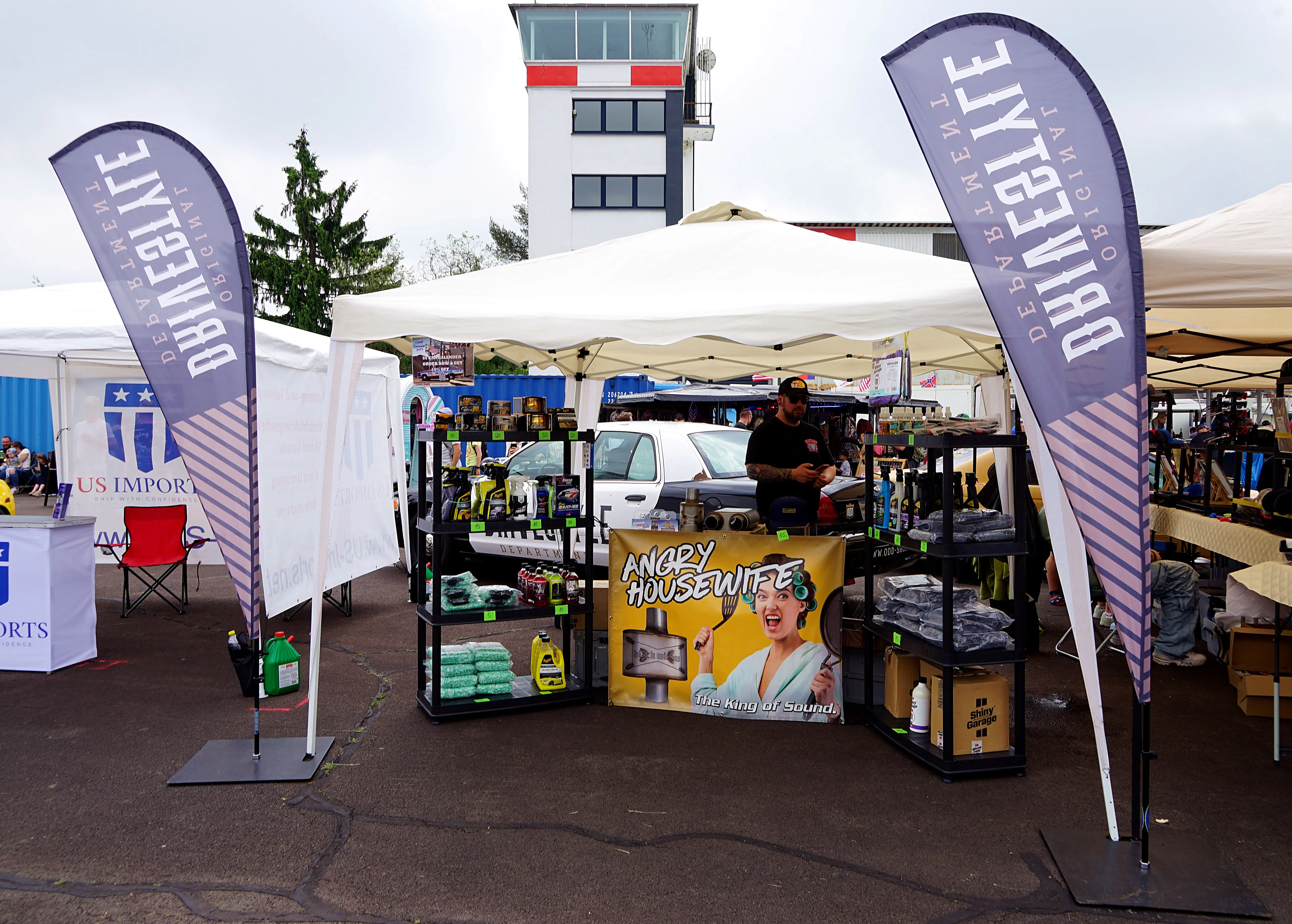 Ein Verkaufsstand auf einem Autotreffen bietet Autopflegeprodukte und Zubehör an. Im Hintergrund ein Kontrollturm und weitere Besucher.