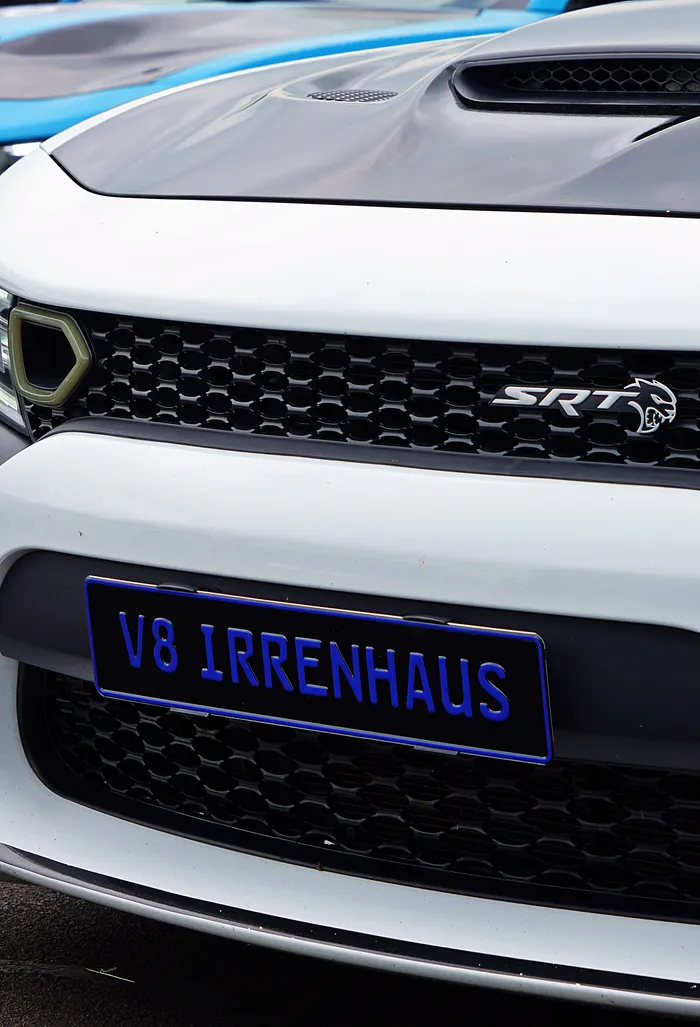 Weißer Sportwagen mit schwarzer Motorhaube und Kühlergrill, auf dem ein blaues Nummernschild mit der Aufschrift „V8 IRRENHAUS“ angebracht ist.