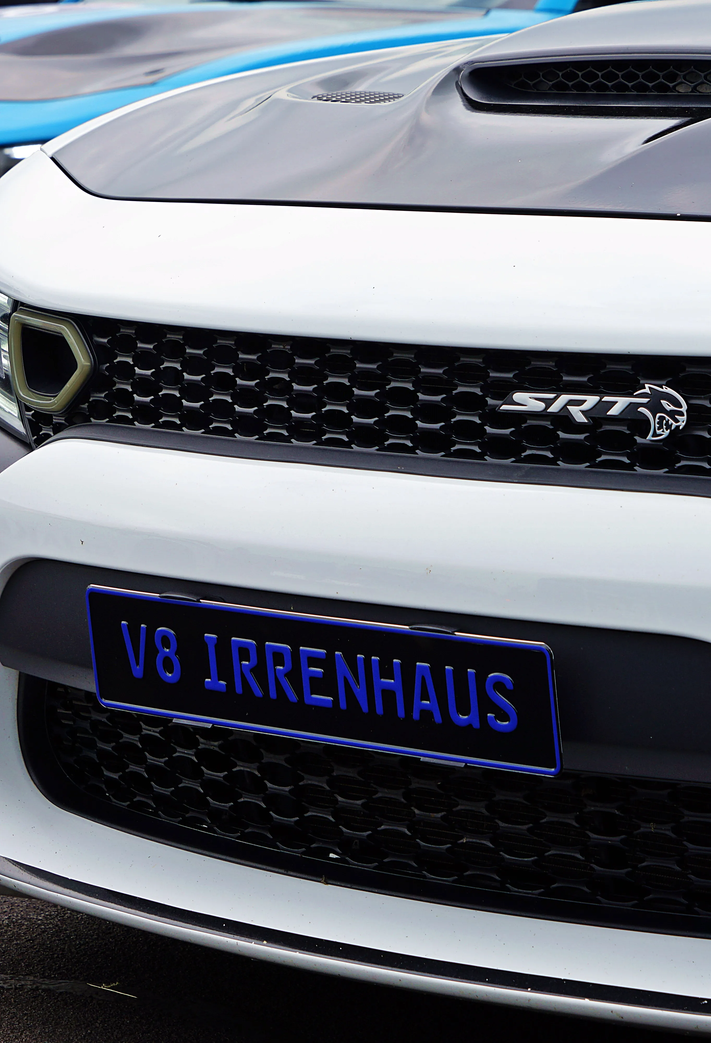 Weißer Sportwagen mit schwarzer Motorhaube und Kühlergrill, auf dem ein blaues Nummernschild mit der Aufschrift „V8 IRRENHAUS“ angebracht ist.