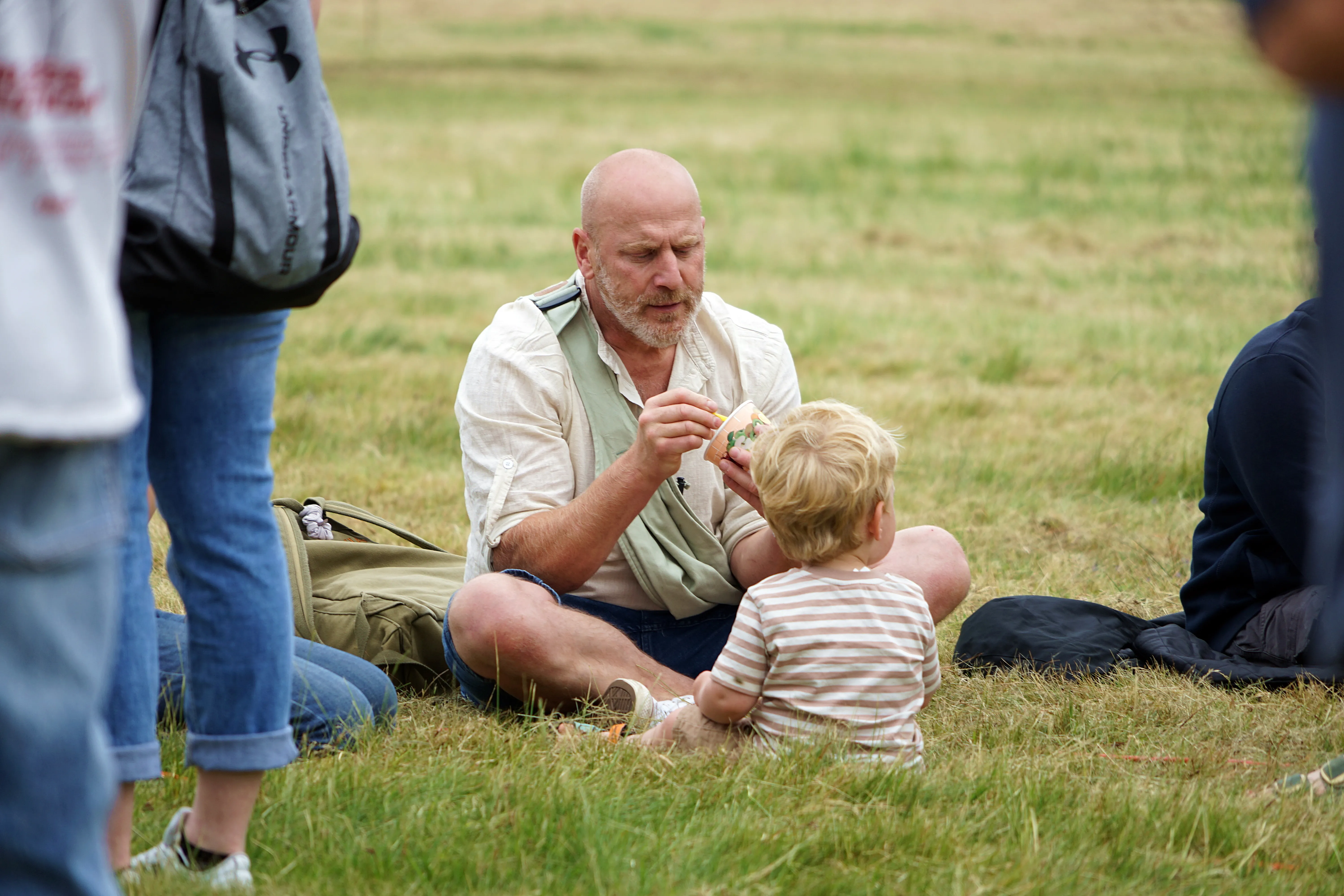 Ein älterer Mann mit Bart sitzt im Gras und füttert ein kleines Kind mit einem Löffel.
