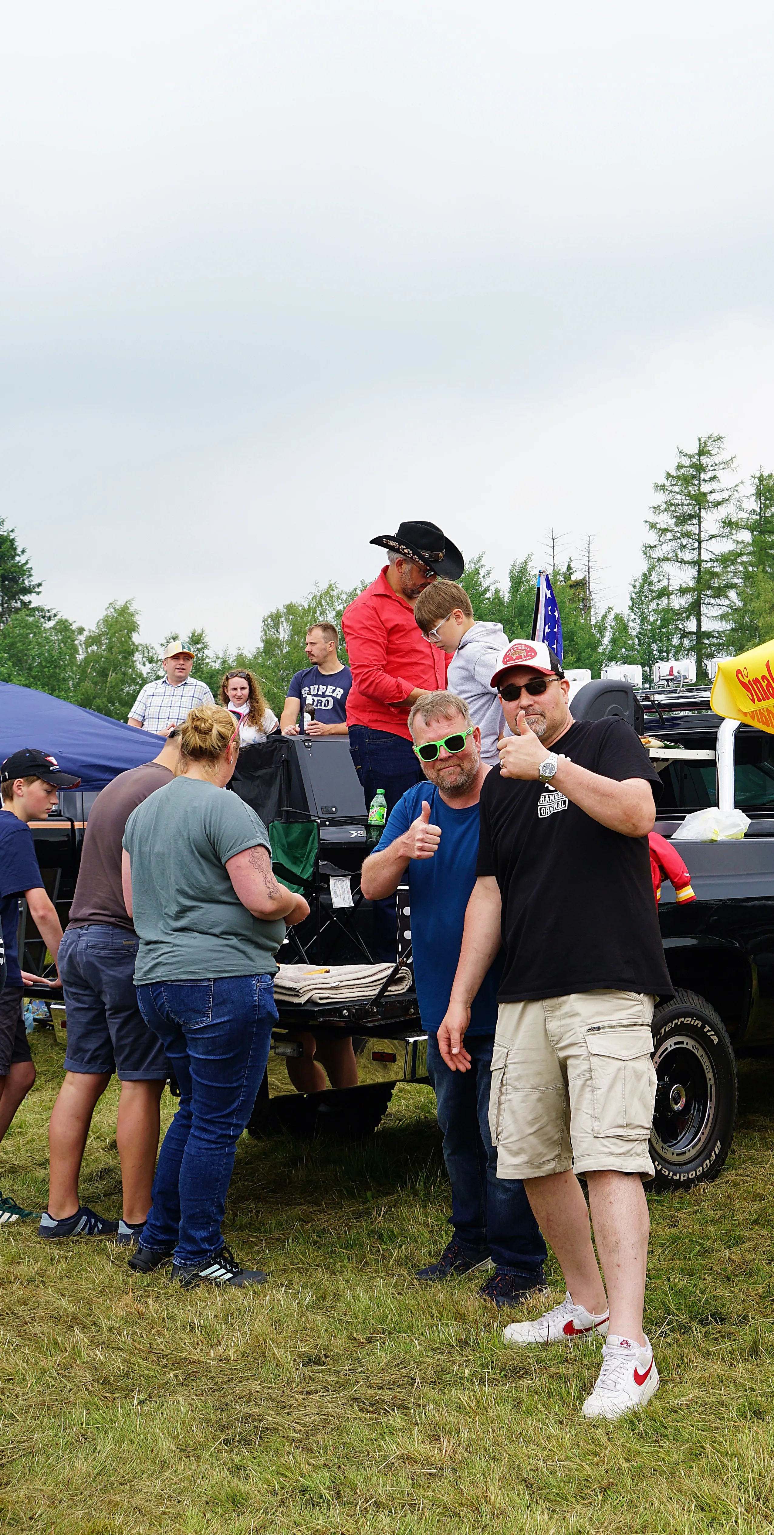 Eine Gruppe von Menschen steht um einen geöffneten Jeep herum, einige unterhalten sich und trinken. Im Hintergrund sind weitere Fahrzeuge und Bäume zu sehen.