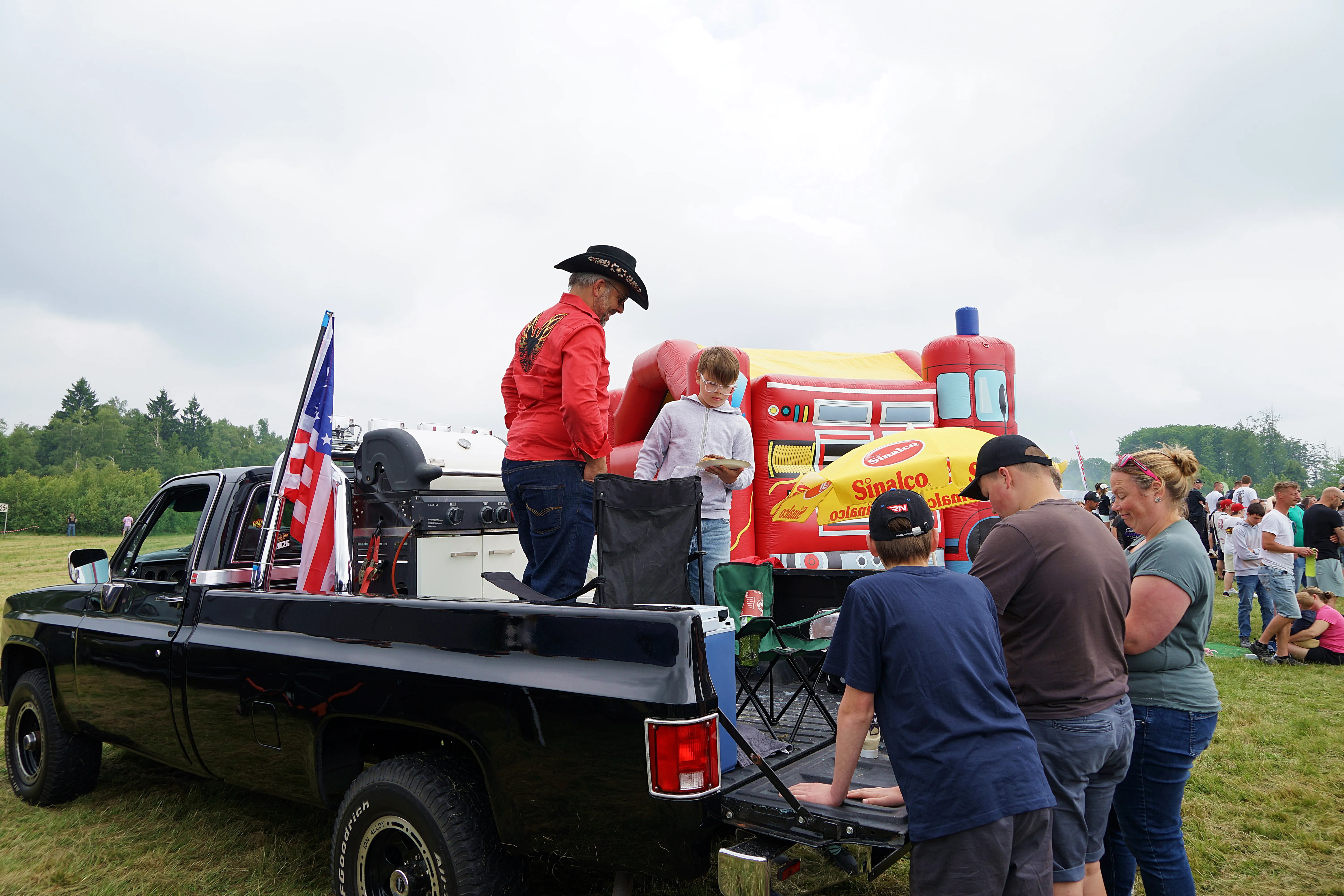 Ein Mann mit Cowboyhut steht auf der Ladefläche eines Pick-up-Trucks, während ein Kind neben ihm steht. Auf der Ladefläche befindet sich ein aufblasbarer Feuerwehrauto und Menschen verteilen sich im Hintergrund.