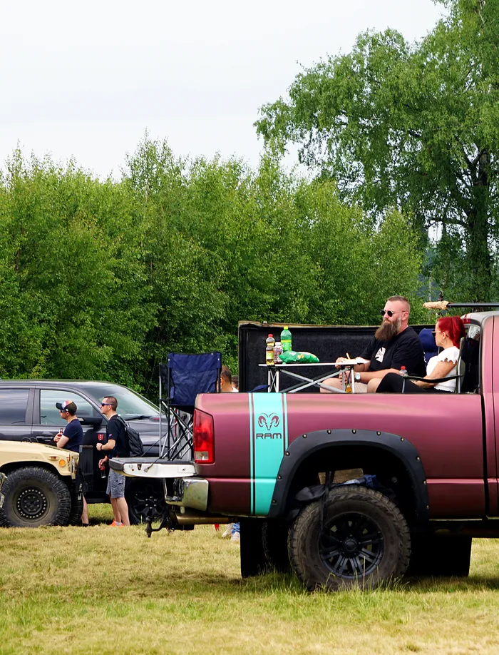Ein dunkelroter Pick-up-Truck mit geöffneter Ladeklappe, in der zwei Personen sitzen und Getränke halten. Im Hintergrund sind weitere Fahrzeuge und Bäume zu sehen.