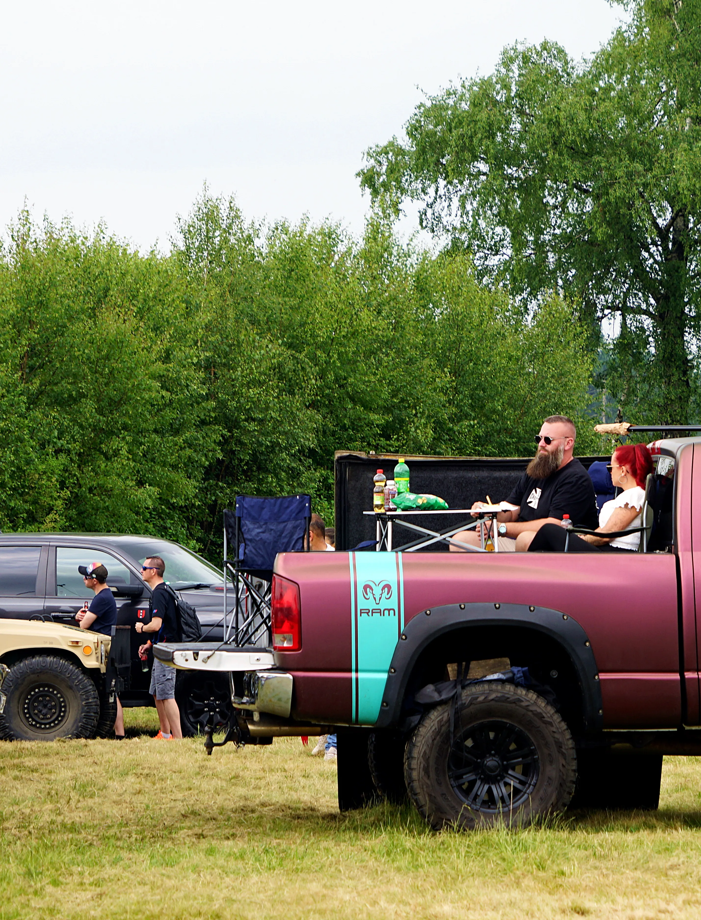 Ein dunkelroter Pick-up-Truck mit geöffneter Ladeklappe, in der zwei Personen sitzen und Getränke halten. Im Hintergrund sind weitere Fahrzeuge und Bäume zu sehen.