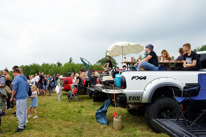 Menschen stehen und sitzen auf und um Pick-up-Trucks auf einer Wiese, einige essen und trinken. Im Hintergrund Bäume und ein bewölkter Himmel.