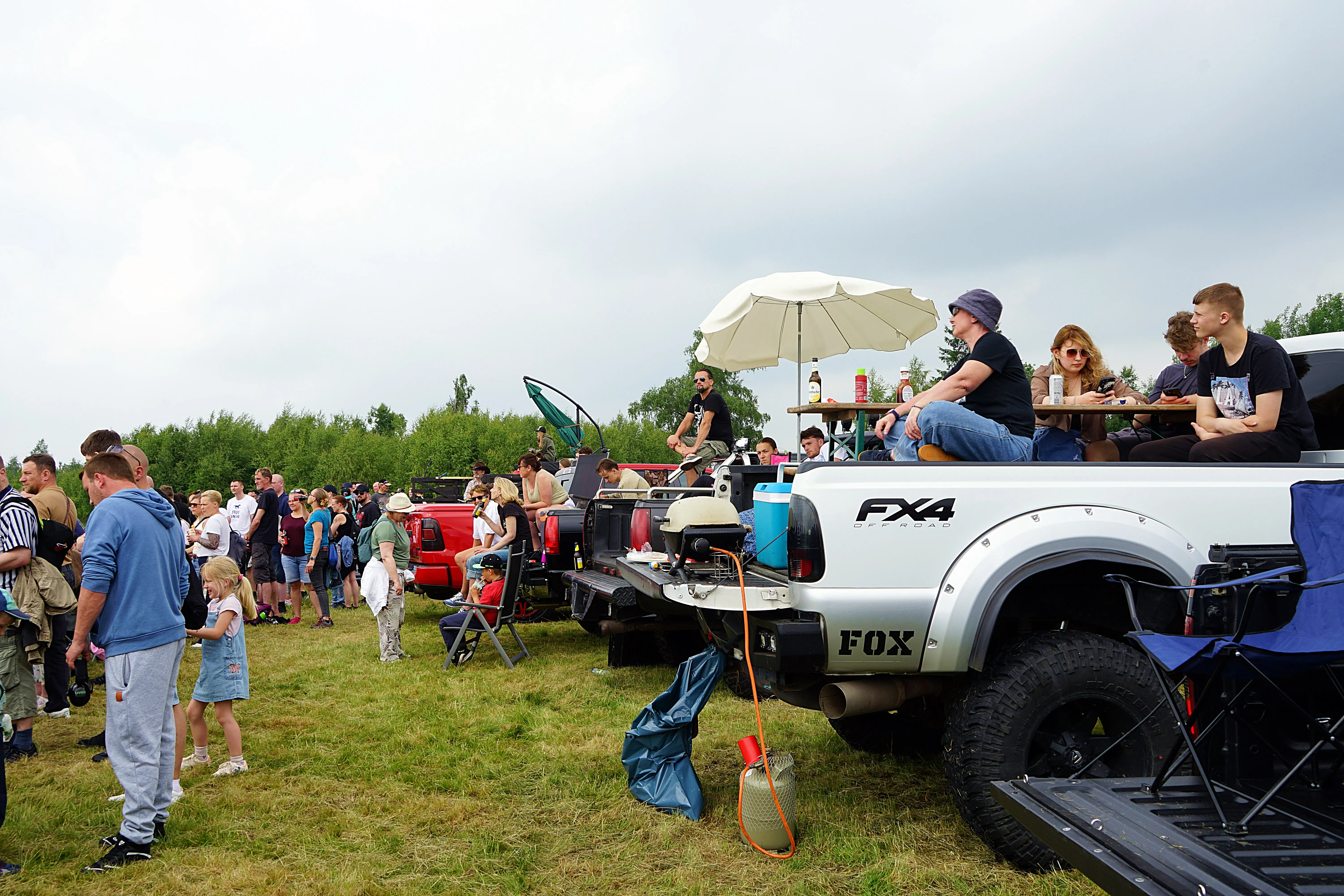 Menschen stehen und sitzen auf und um Pick-up-Trucks auf einer Wiese, einige essen und trinken. Im Hintergrund Bäume und ein bewölkter Himmel.