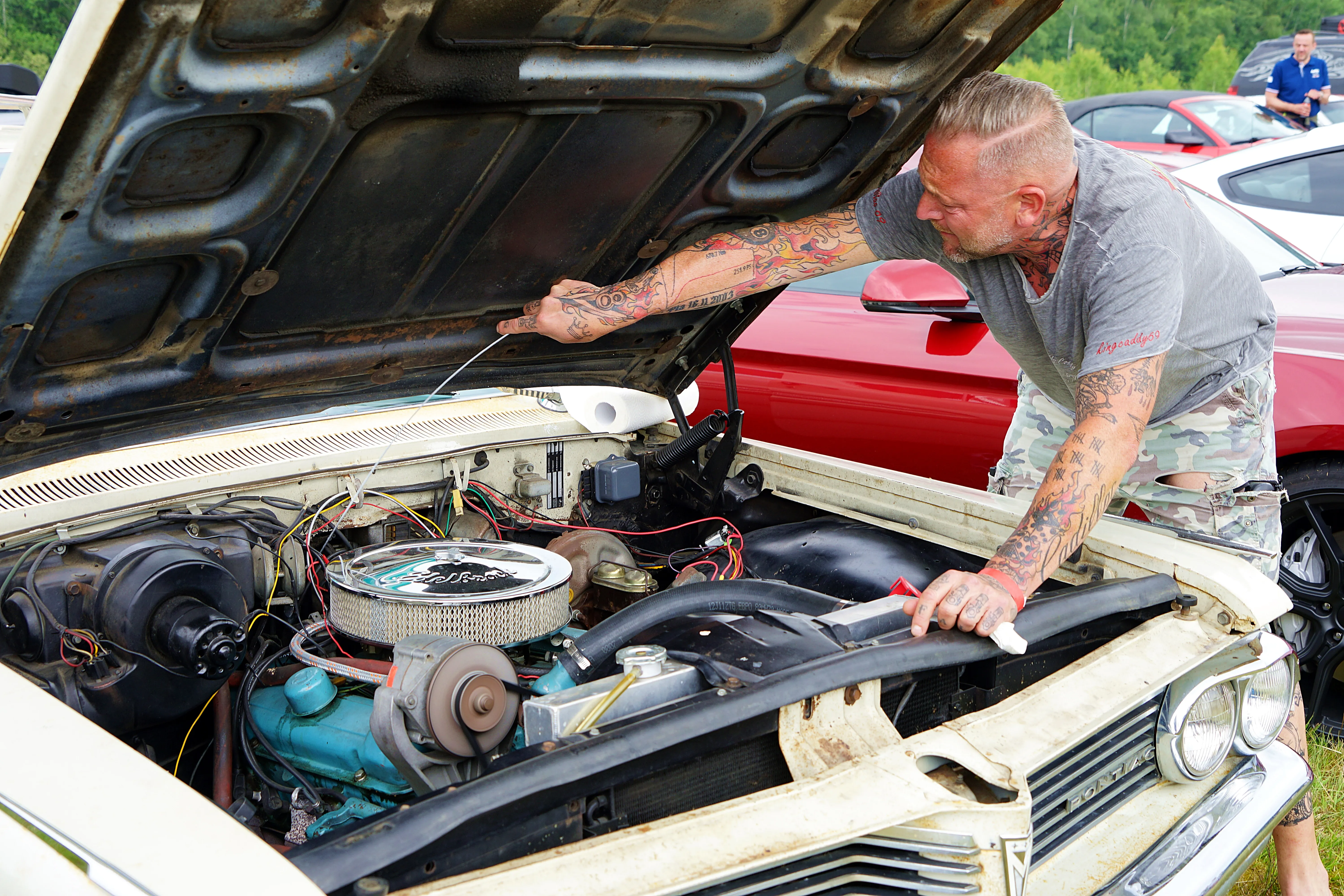 Ein Mann mit Tattoos arbeitet an einem geöffneten Motorraum eines klassischen weißen Autos. Er hält ein Werkzeug in der Hand und inspiziert den Motor.
