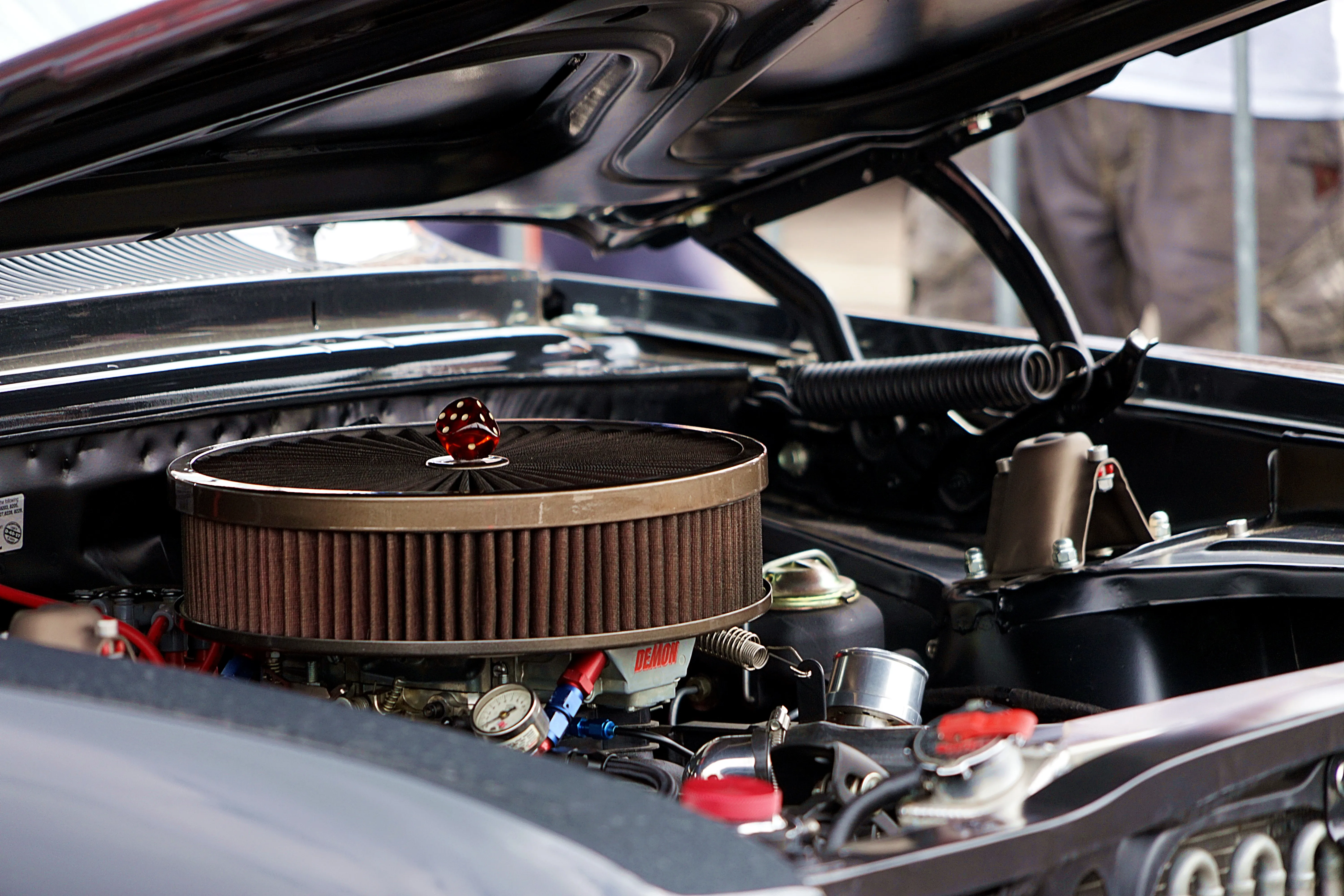 Detailaufnahme des Motors eines Oldtimers mit einem großen, runden Luftfilter und roten Würfeln als Dekoration.
