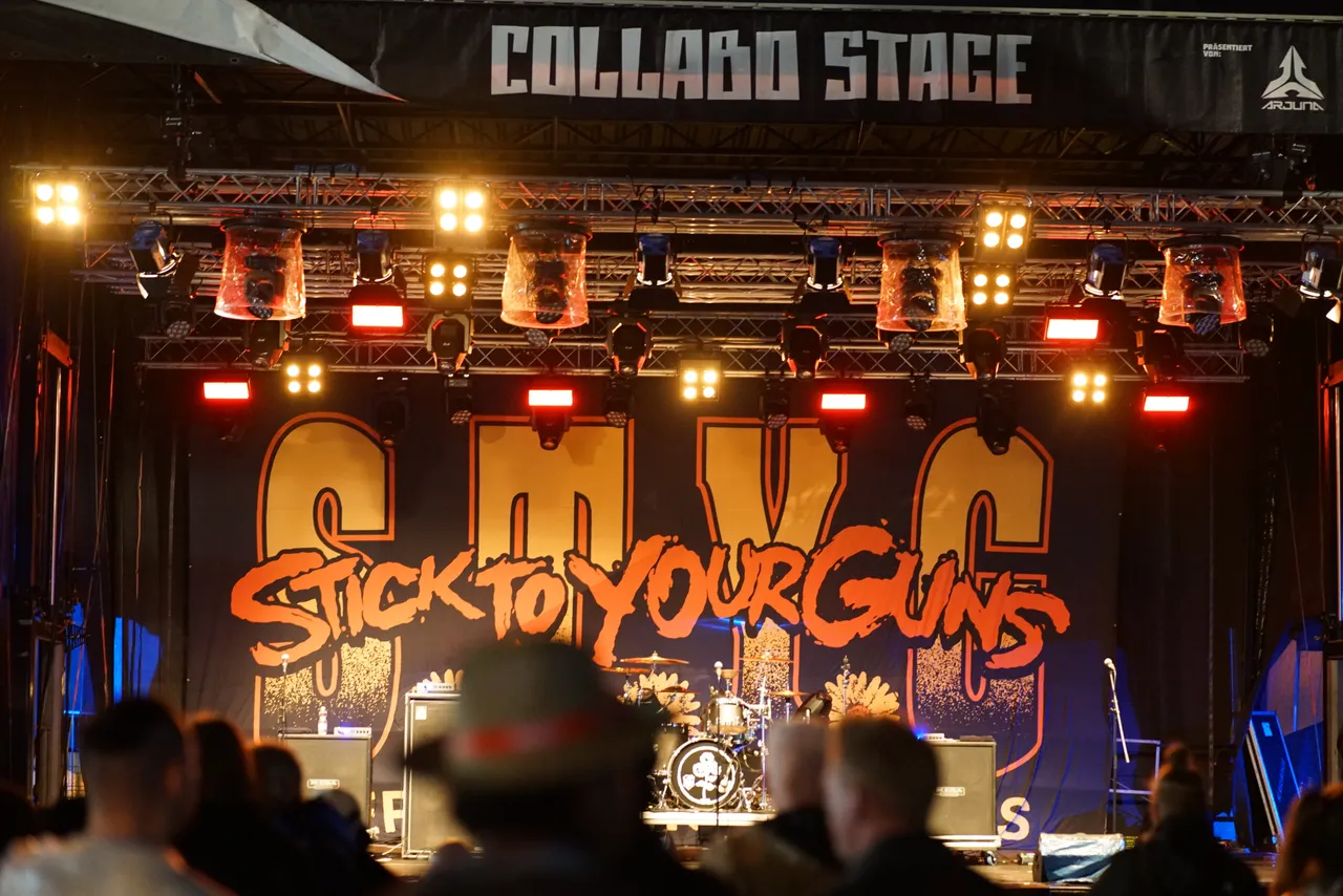 Die Collabo Stage mit dem großen orangefarbenen Schriftzug der Band Stick To Your Guns und farbiger Bühnenbeleuchtung vor einem Publikum.