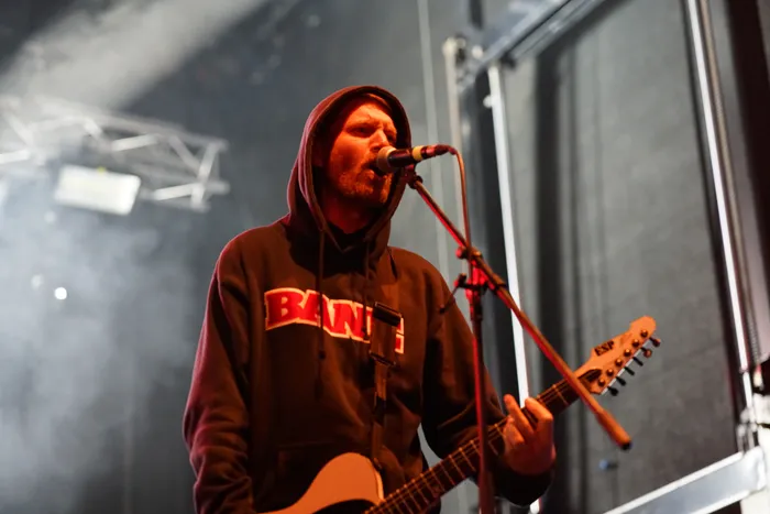 Ein bärtiger Musiker der Band Stick To Your Guns singt in ein Mikrofon und spielt E-Gitarre auf einer Bühne.