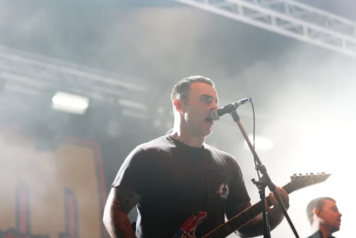 Ein Sänger der Band Stick To Your Guns singt in ein Mikrofon und spielt Gitarre auf einer nebligen Bühne.