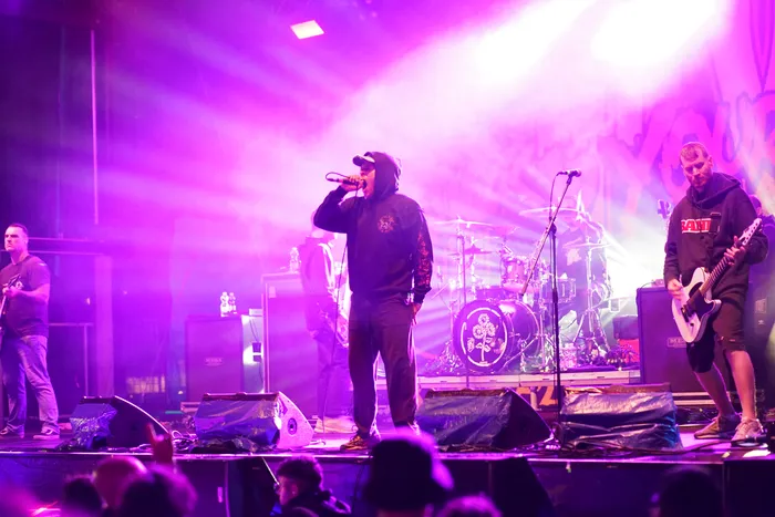 Die Band Stick To Your Guns tritt live auf einer Bühne unter intensiver violetter Beleuchtung auf. Im Zentrum singt der Sänger, rechts spielt ein Gitarrist.