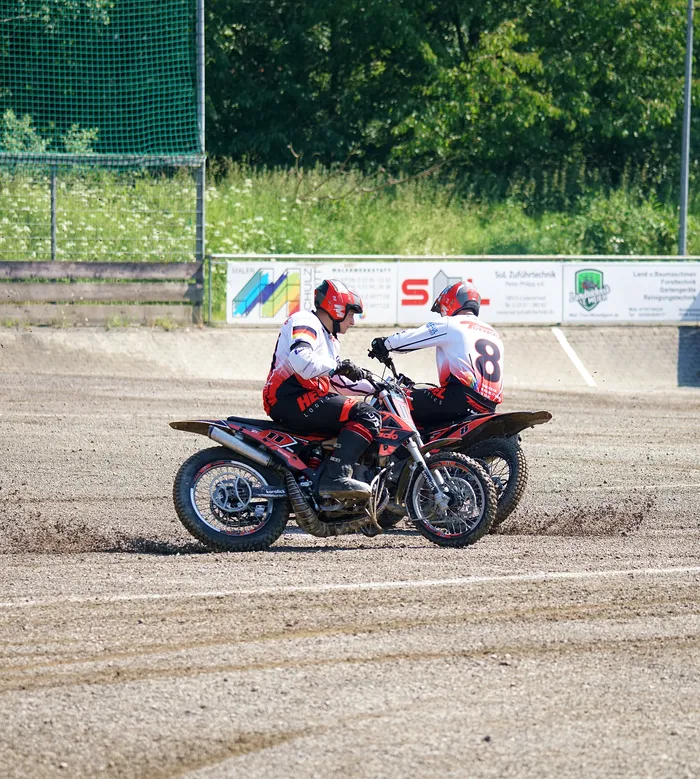 Zwei Motorradfahrer mit den Nummern 8 und 11 fahren auf einer staubigen Rennstrecke nebeneinander. Im Hintergrund eine grüne Hecke und Werbebanner.