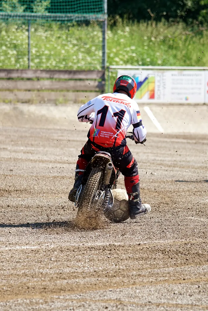 Ein Motoballspieler mit der Nummer 11 auf dem roten Trikot fährt auf einem staubigen Platz mit einem Motorrad. Im Hintergrund ist eine grüne Hecke und ein Gebäude zu sehen.