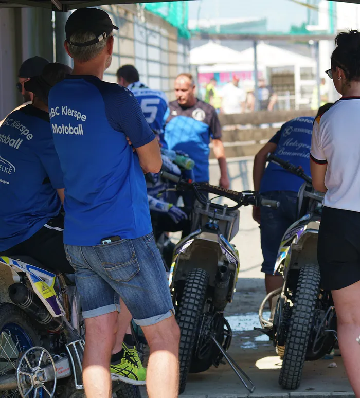 Ein Mann mit blauem T-Shirt und Jeansshorts steht mit dem Rücken zum Betrachter vor einem Motoball-Motorrad. Im Hintergrund weitere Personen und Motorräder in einer Art Fahrerlager.