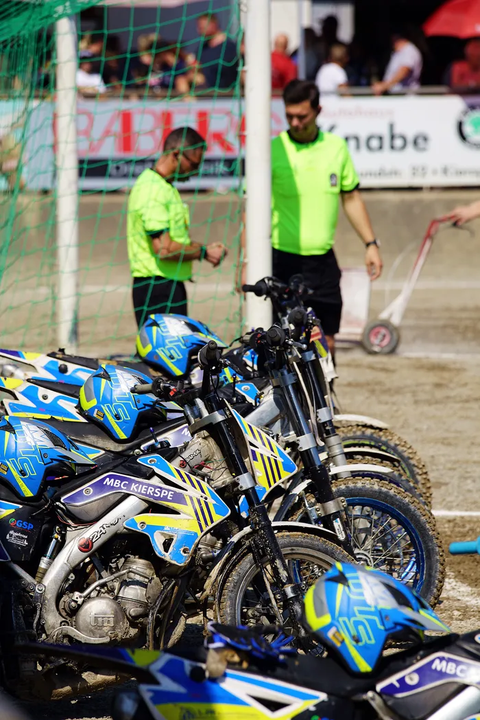 Mehrere Motoball-Motorräder stehen nebeneinander, bereit für das Spiel. Im Hintergrund stehen zwei Offizielle in grünen Hemden und beobachten die Vorbereitungen.