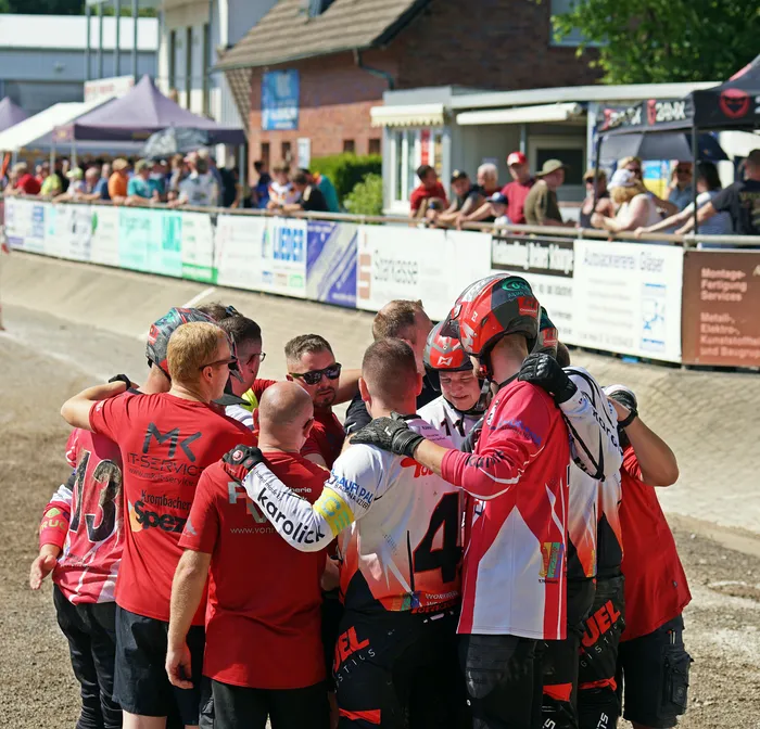 Eine Gruppe von Motoballspielern in roten Trikots steht eng zusammen und feiert, einige mit erhobenen Armen. Im Hintergrund sind Zuschauer und die Motoballbahn zu sehen.