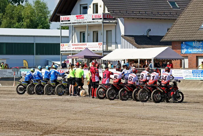 Motoballspieler auf Motorrädern stehen bereit für den Spielbeginn auf einer staubigen Bahn, umgeben von Zuschauern und Werbebannern.