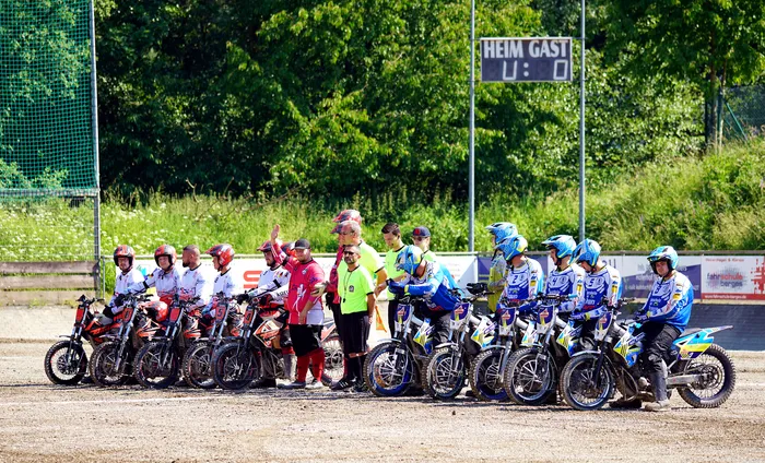 Motoballspieler stehen mit ihren Motorrädern auf einer Sandbahn bereit, ein Schiedsrichter gestikuliert. Im Hintergrund eine Anzeigetafel mit dem Stand 0:0.