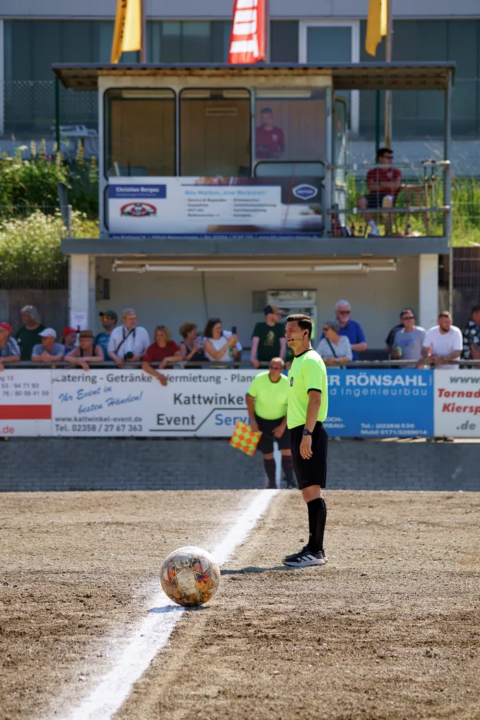 Ein Schiedsrichter in schwarzer Hose und gelbem Trikot steht auf einem Sandplatz neben einem Motoball. Im Hintergrund ein Banner mit Werbeaufdruck und Zuschauer.