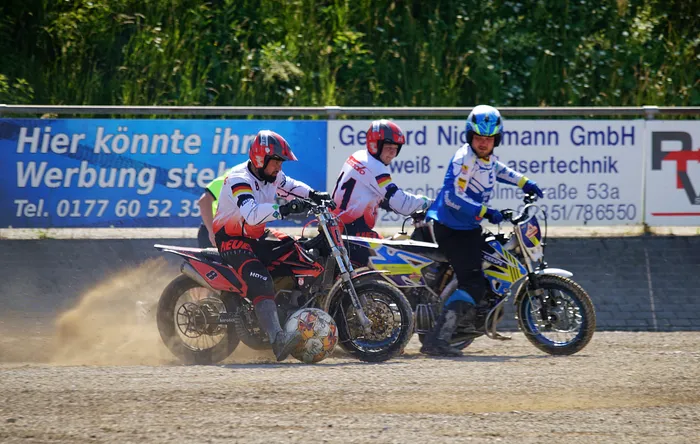Drei Motoballspieler auf Motorrädern rasen auf einer staubigen Bahn nebeneinander. Im Hintergrund eine Werbebande und Bäume.