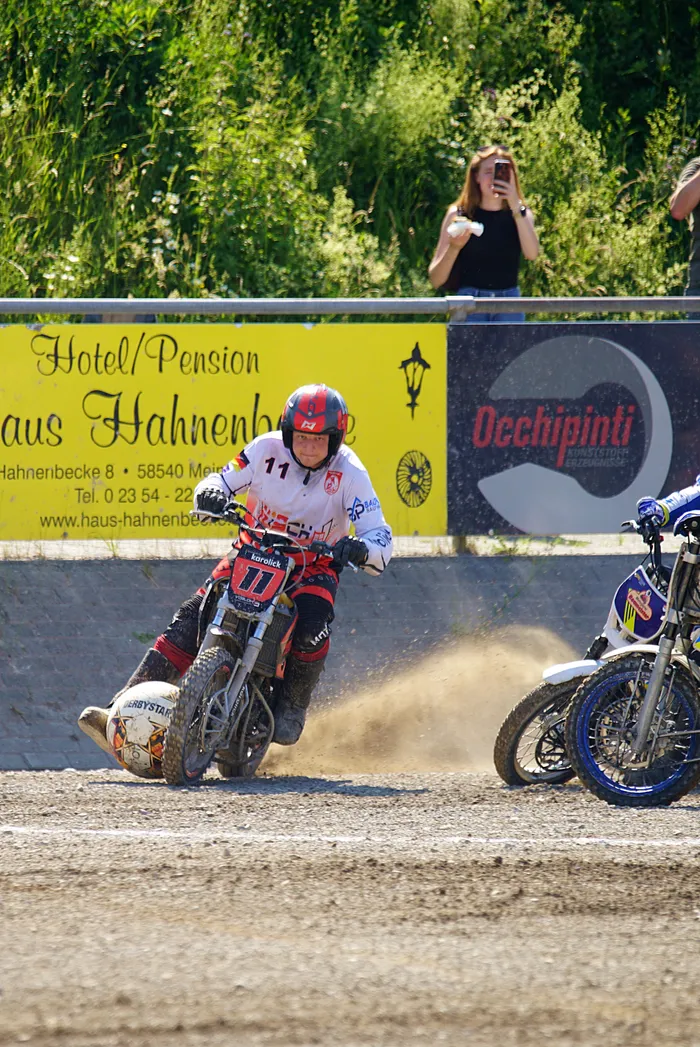 Ein Motoballspieler mit der Startnummer 11 fährt auf einem Motorrad auf einer staubigen Rennstrecke, während ein weiterer Fahrer im Hintergrund folgt. Im Hintergrund ist ein Werbebanner für ein Hotel/Pension und ein Restaurant zu sehen.