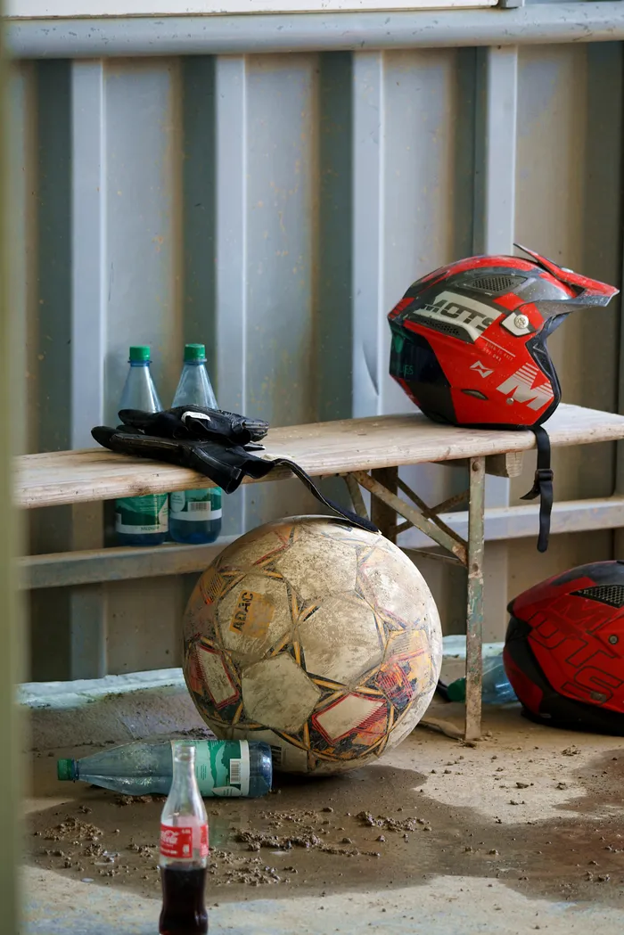 Ein roter Motocross-Helm, ein Fußball und mehrere Glasflaschen stehen auf einer Holzbank vor einer metallenen Wand. Der Fußball ist abgenutzt und weist Gebrauchsspuren auf.