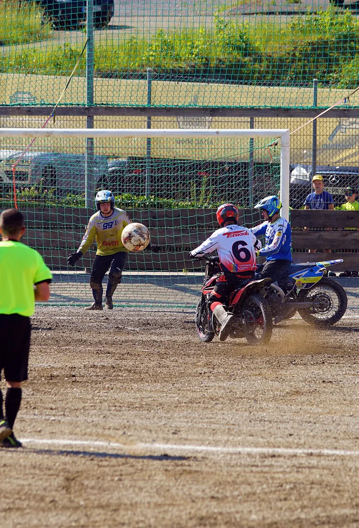 Ein Motoballspieler mit der Nummer 6 auf einem Motorrad fährt auf das Tor zu, während ein Torwart den Ball abwehrt. Im Hintergrund stehen weitere Spieler und Zuschauer.