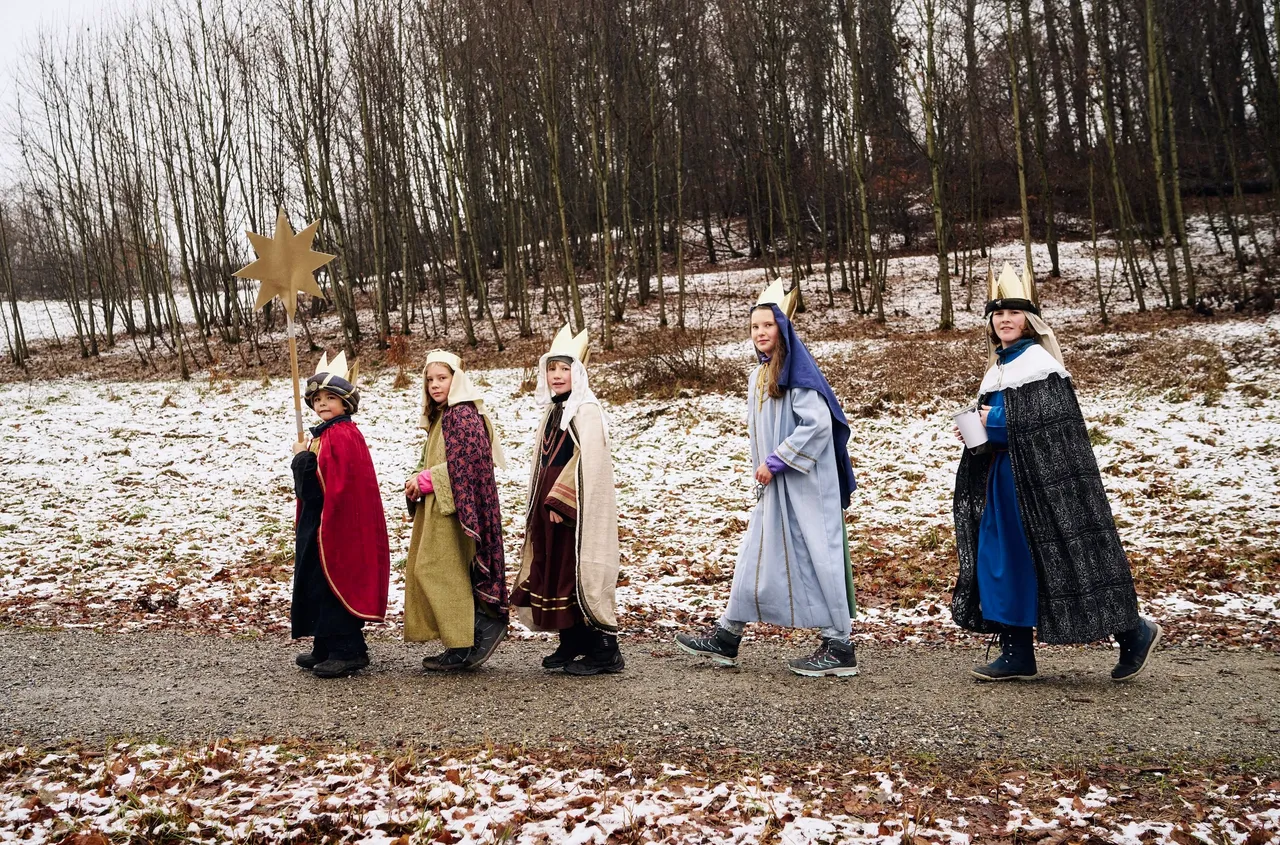 Fünf Kinder in Sternsinger-Kostümen mit Kronen und Umhängen gehen in einer Reihe über einen verschneiten Waldweg.