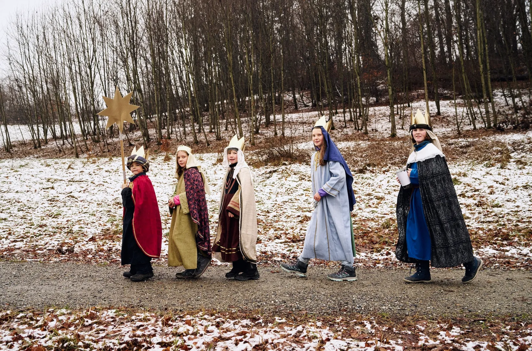 Fünf Kinder in Sternsinger-Kostümen mit Kronen und Umhängen gehen in einer Reihe über einen verschneiten Waldweg.