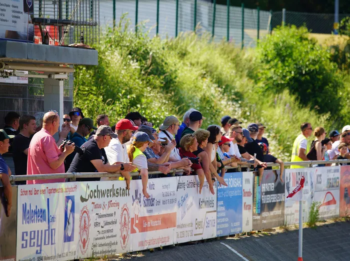 Zuschauer stehen an einer Absperrung und beobachten ein Motoball-Spiel. Werbebanner für lokale Unternehmen sind entlang der Absperrung angebracht.